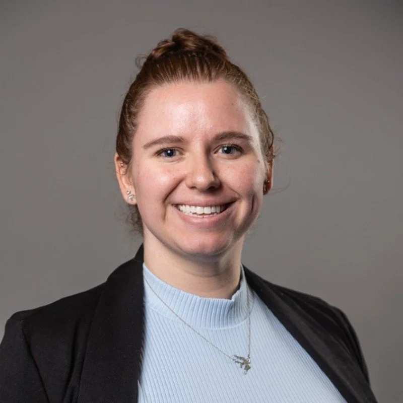 Brianna Hnath, a portrait of a young woman with red hair tied back, wearing a black blazer over a light blue turtleneck sweater, smiling against a plain gray background.
