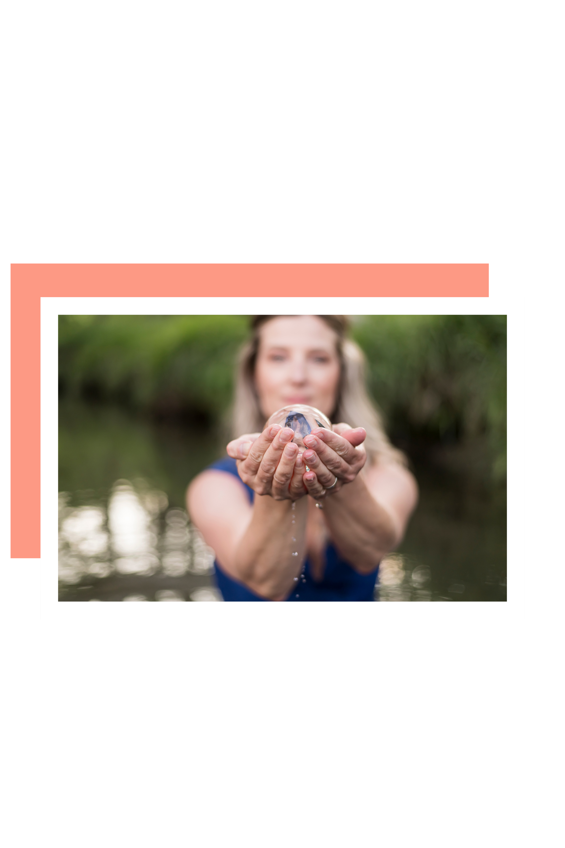 A woman in a blue top holding a shiny seashell towards the camera with water dripping from it, outdoors with blurred green foliage background.