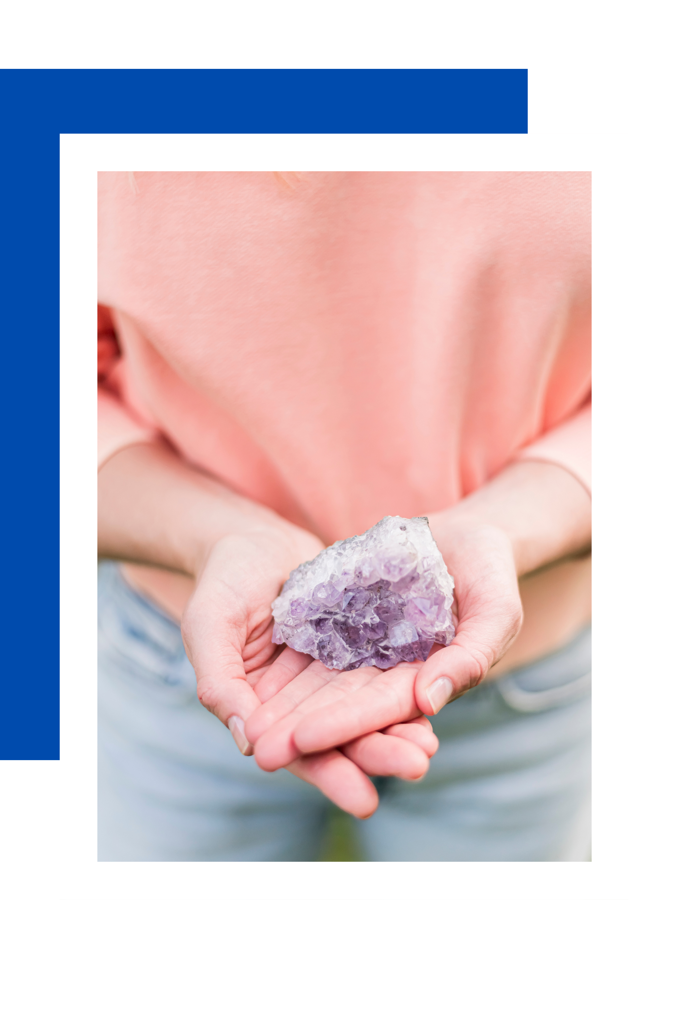 Person holding a purple amethyst crystal in both hands, wearing a light pink shirt and light-colored jeans, against a blurred green background.