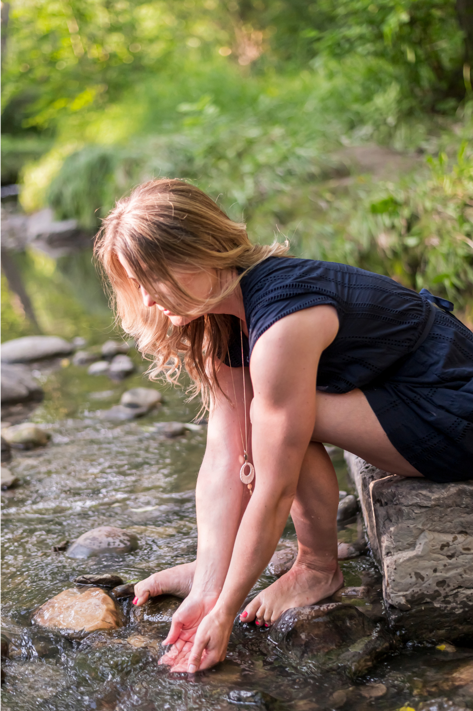 A woman with long, wavy hair, wearing a black dress and a pendant necklace, crouching by a rocky stream, touching the water with her hands and feet, surrounded by lush green foliage.