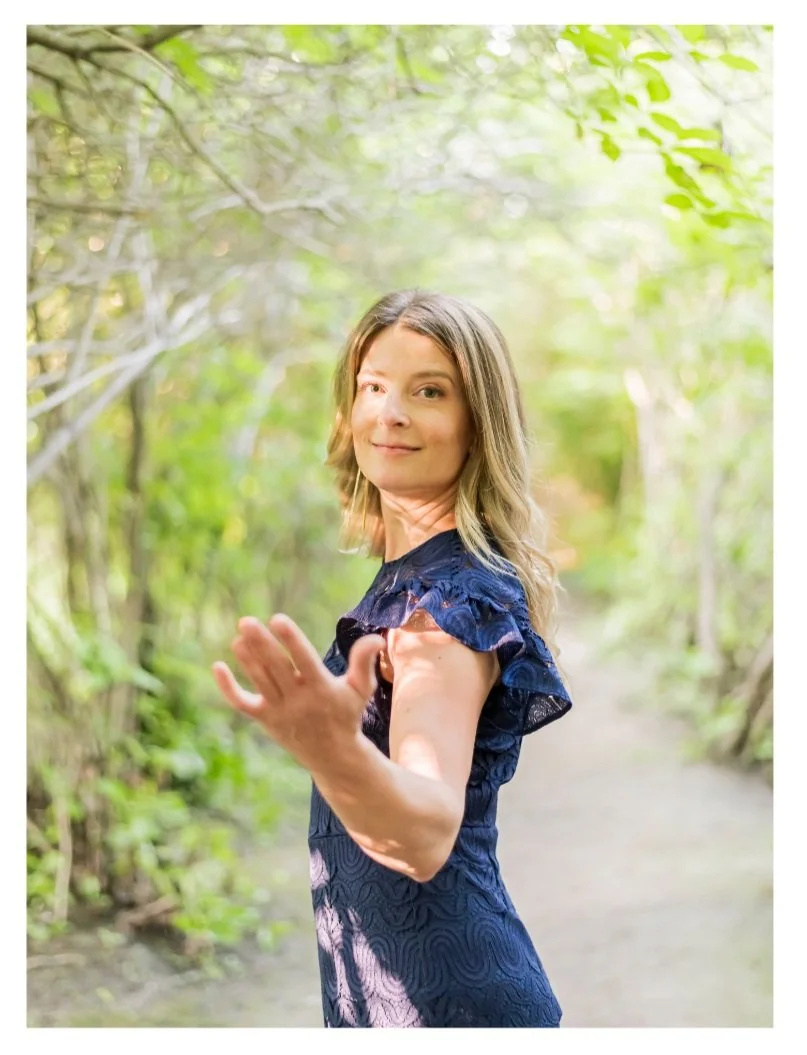 Woman in a blue dress standing on a nature trail surrounded by green trees, reaching out her hand towards the camera.