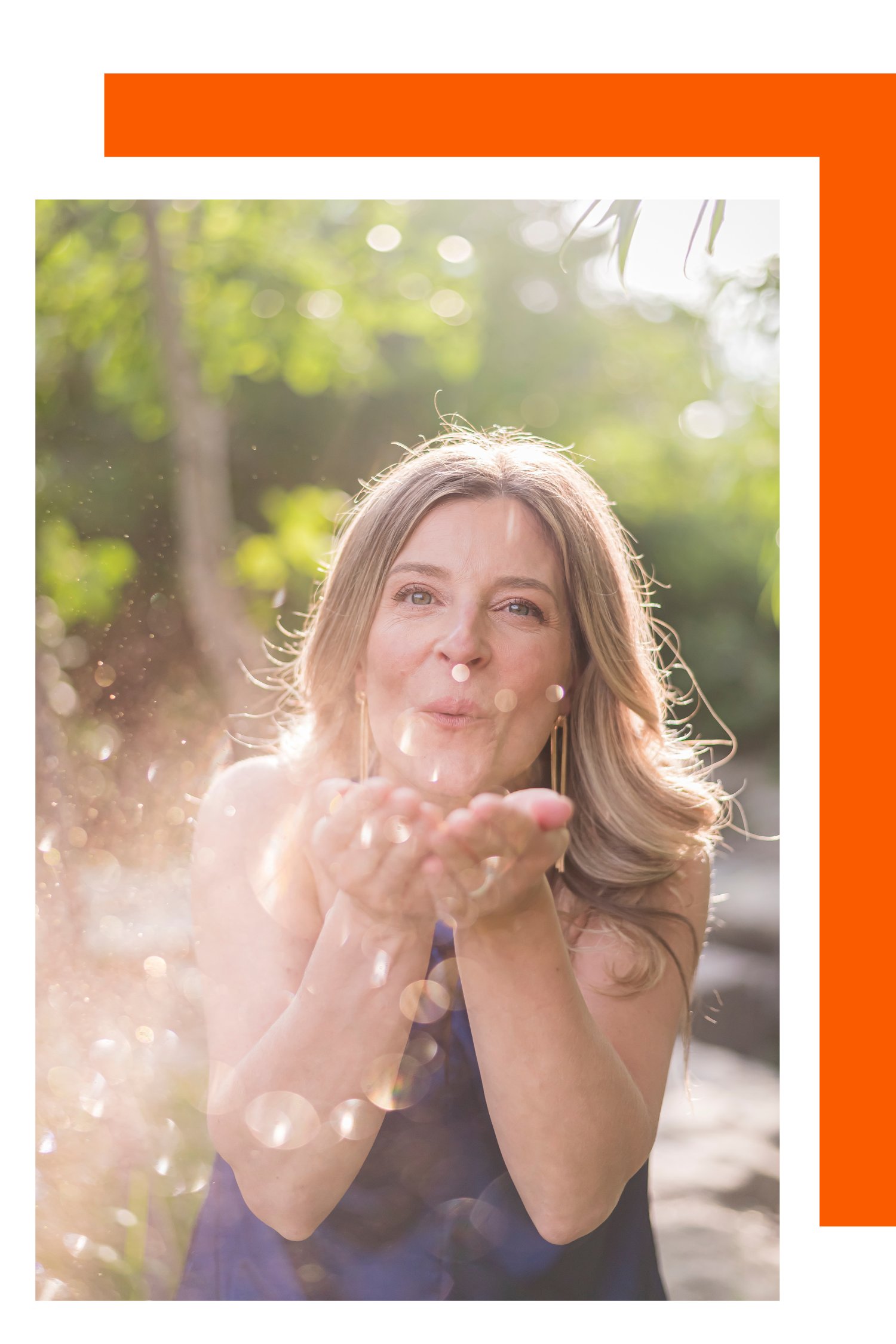 Woman in an outdoor setting blowing glitter or sparkles from her hands, with sunlight filtering through the trees behind her.