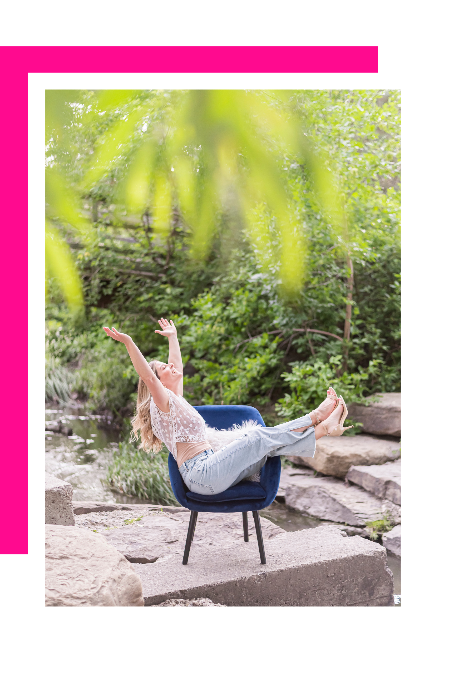 A young woman with long blonde hair is sitting on a blue chair outdoors near rocks and a small stream, with arms raised and head tilted back, surrounded by green foliage.