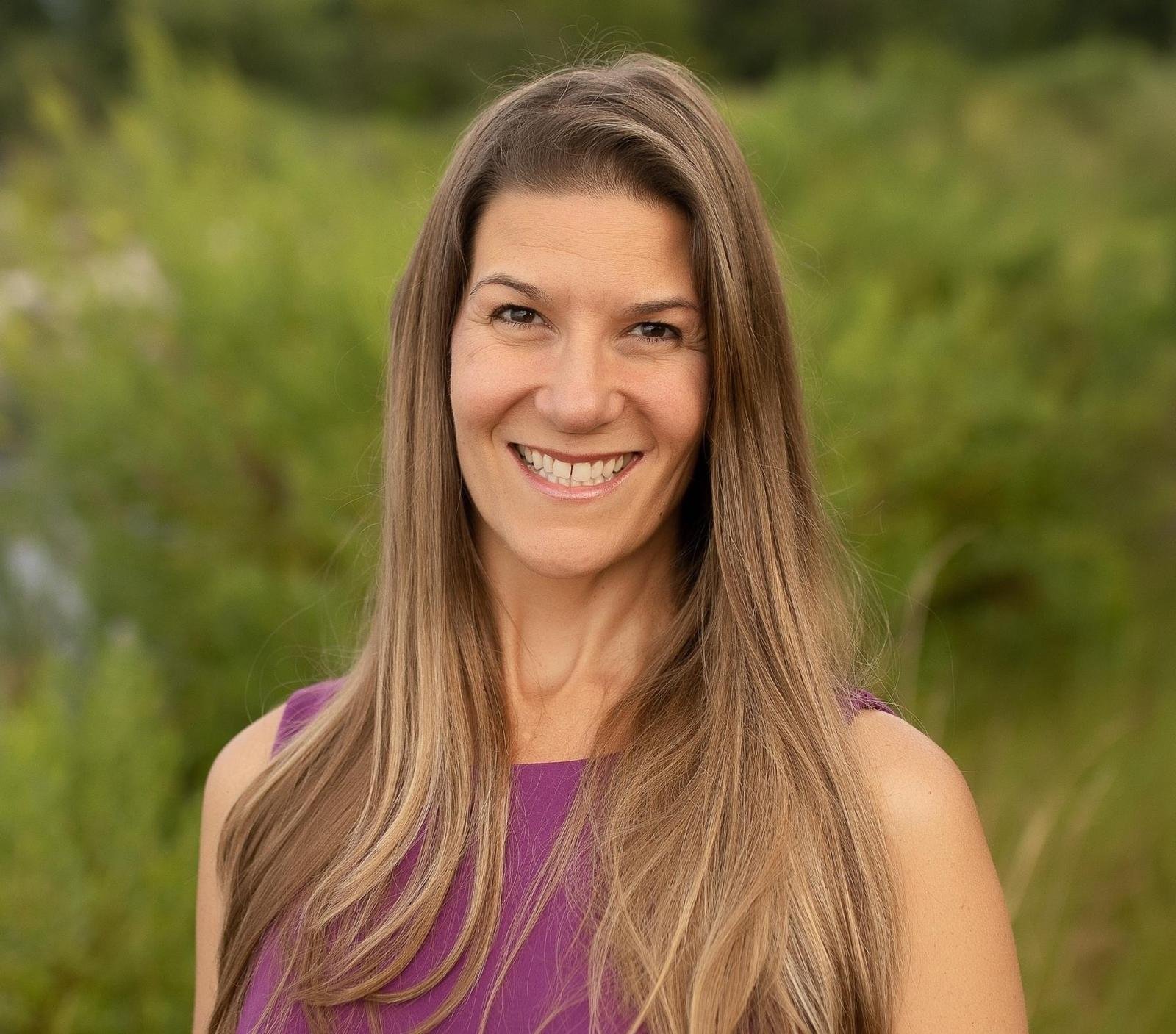 A woman with long, light brown hair smiling outdoors with a blurred green forest background.