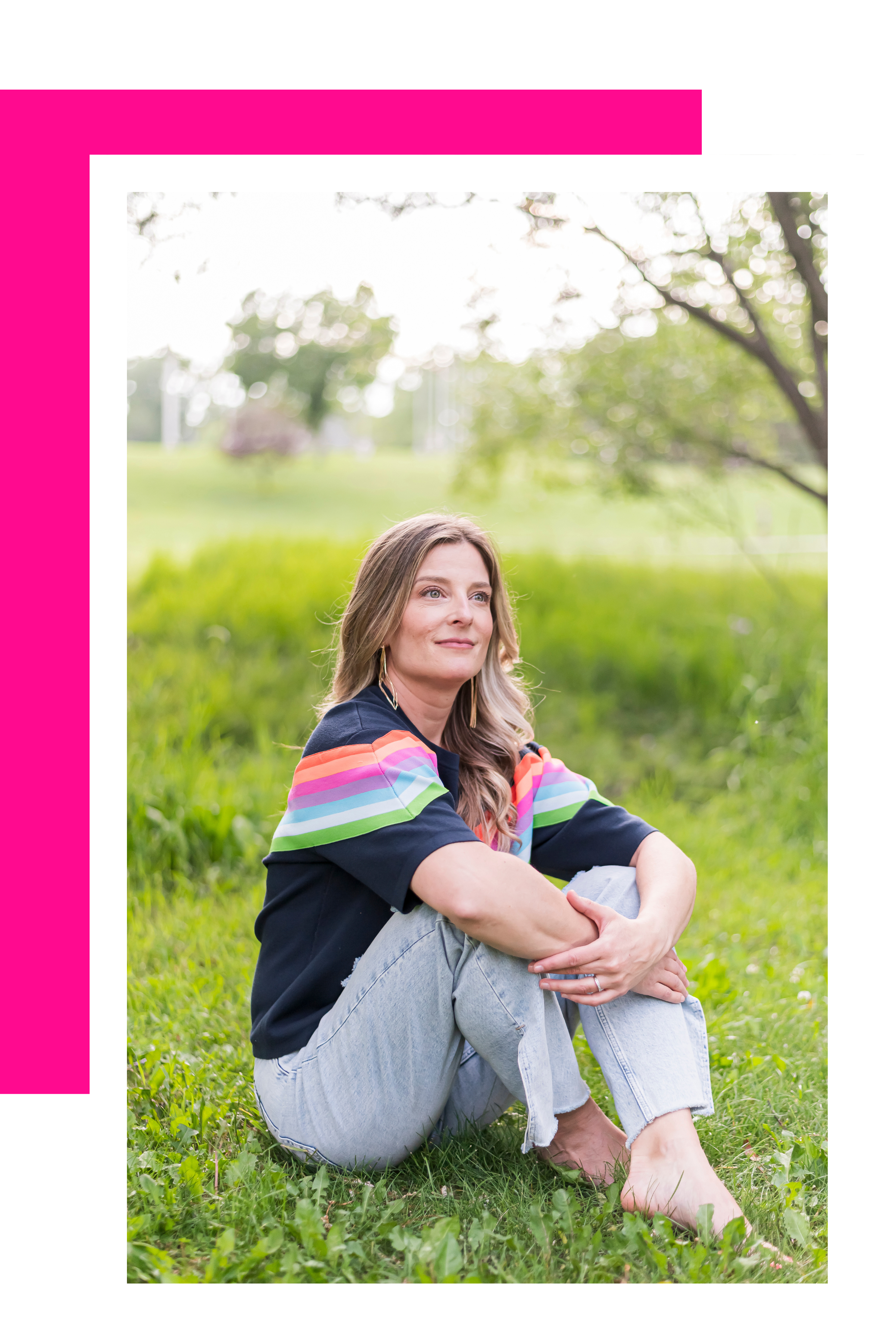 A woman sitting on the grass in a park during daytime, with green trees and a blurred background, wearing a black shirt with rainbow-colored stripes on the shoulders and light blue jeans.