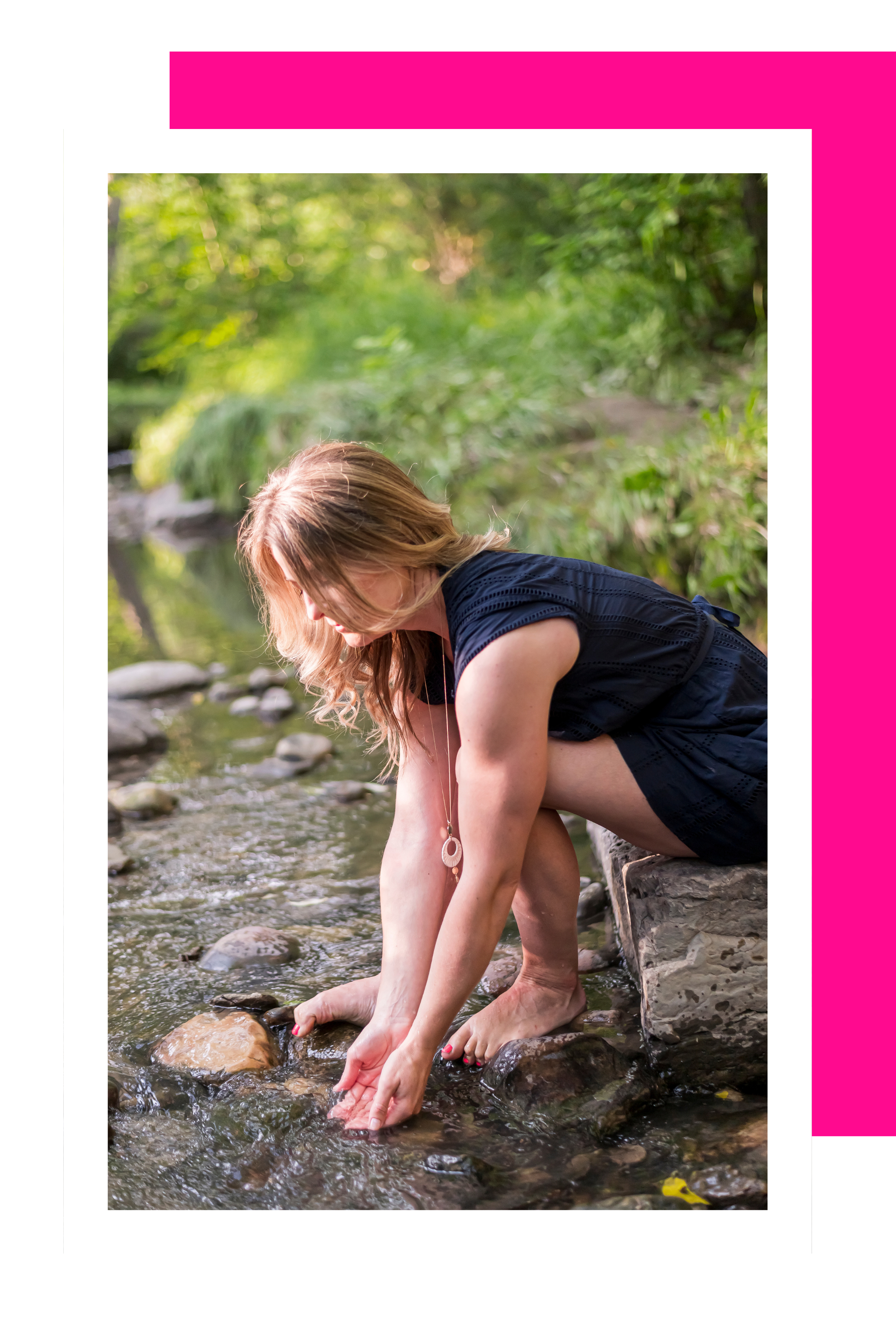 A woman sitting on a rock by a river, leaning forward and touching the water with her hands, surrounded by lush greenery.