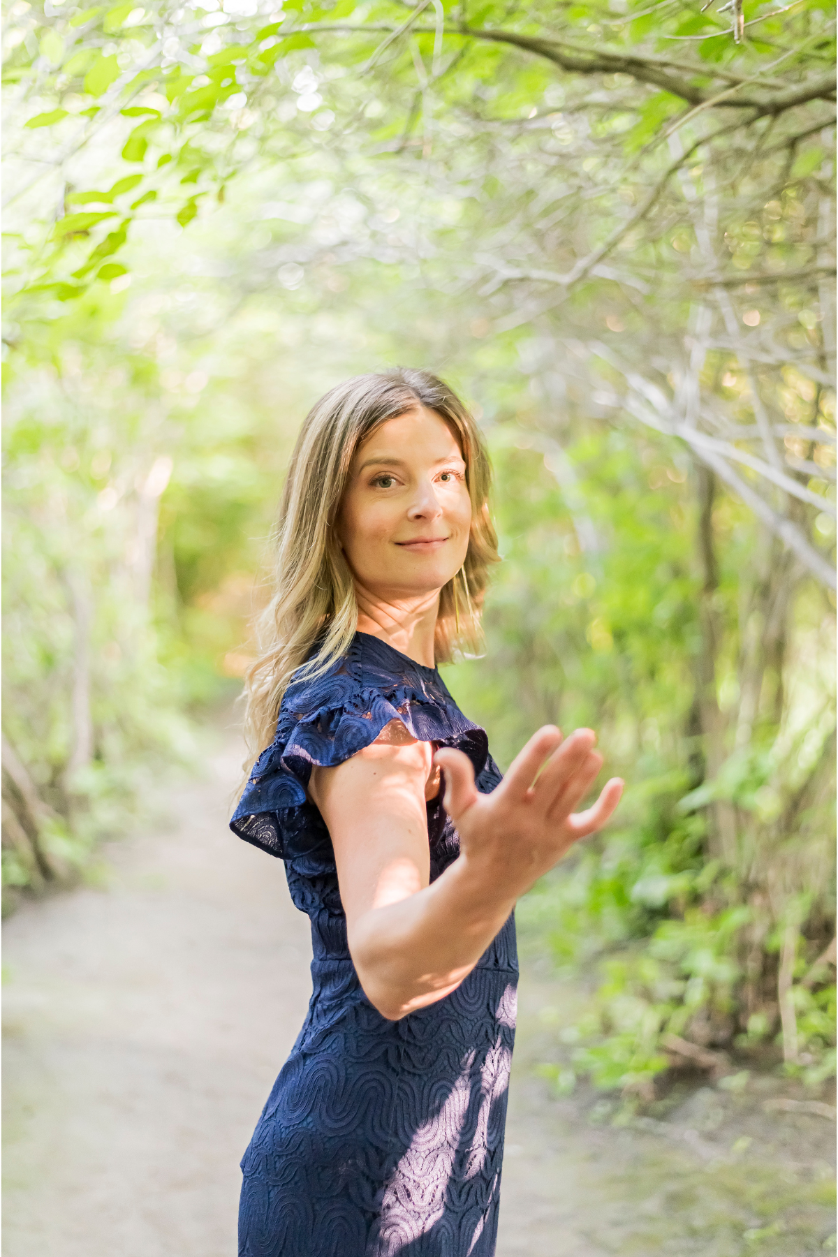 A woman in a blue dress with ruffled sleeves standing on a leafy path in a forest, looking at the camera and extending her arm forward with an open hand.
