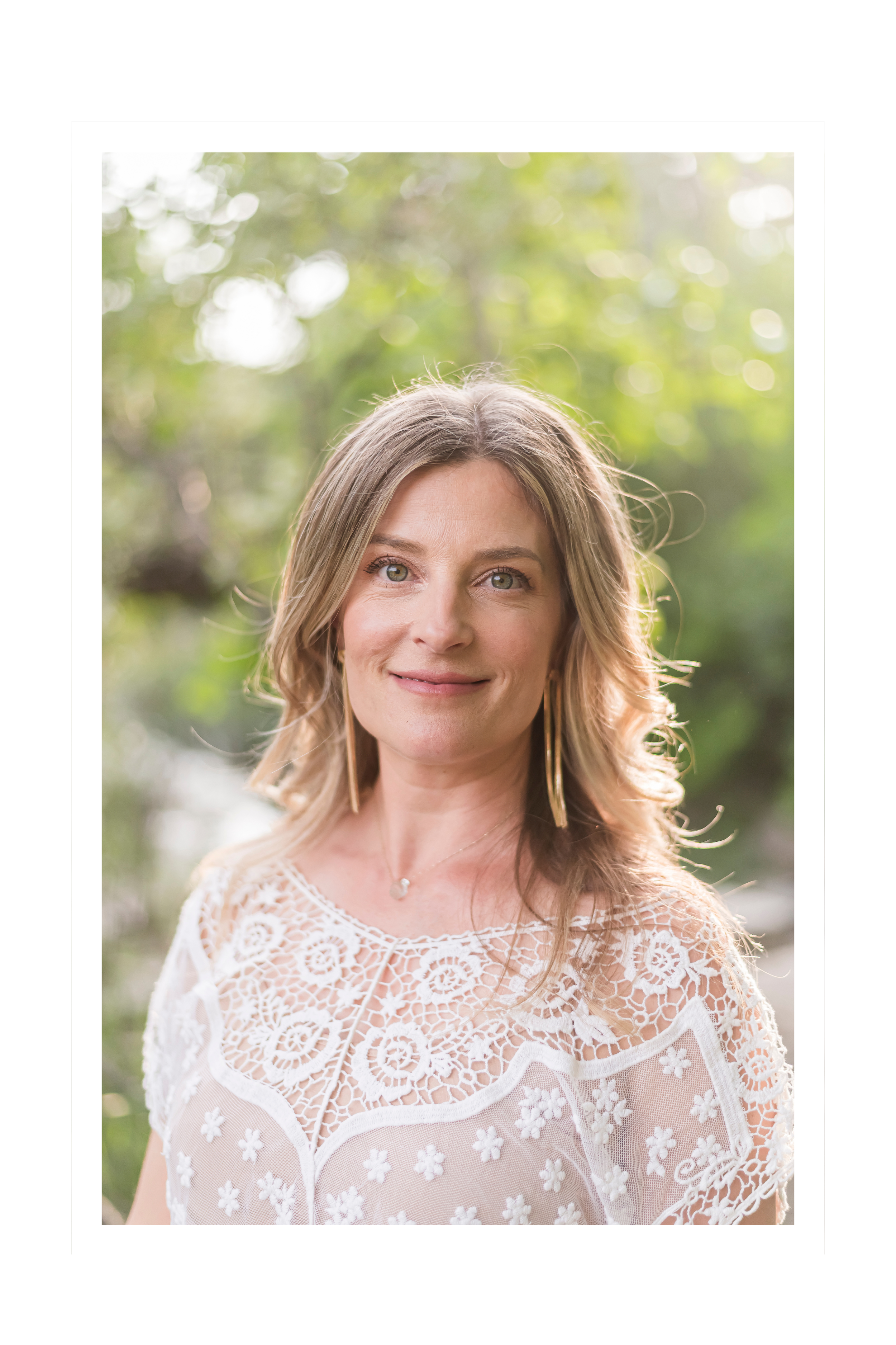 A woman with shoulder-length wavy blonde hair smiling outdoors with blurred green trees in the background, wearing a white embroidered lace top and gold earrings.