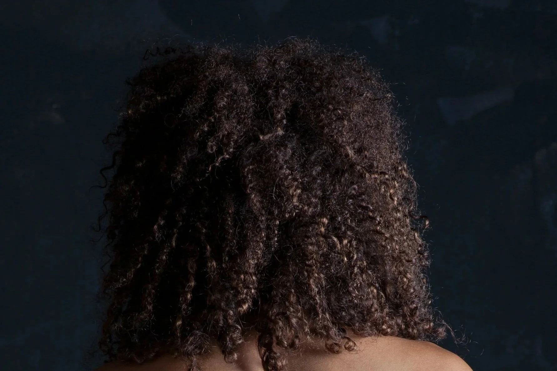 Back of a person's head with natural curly, textured hair against a dark background.