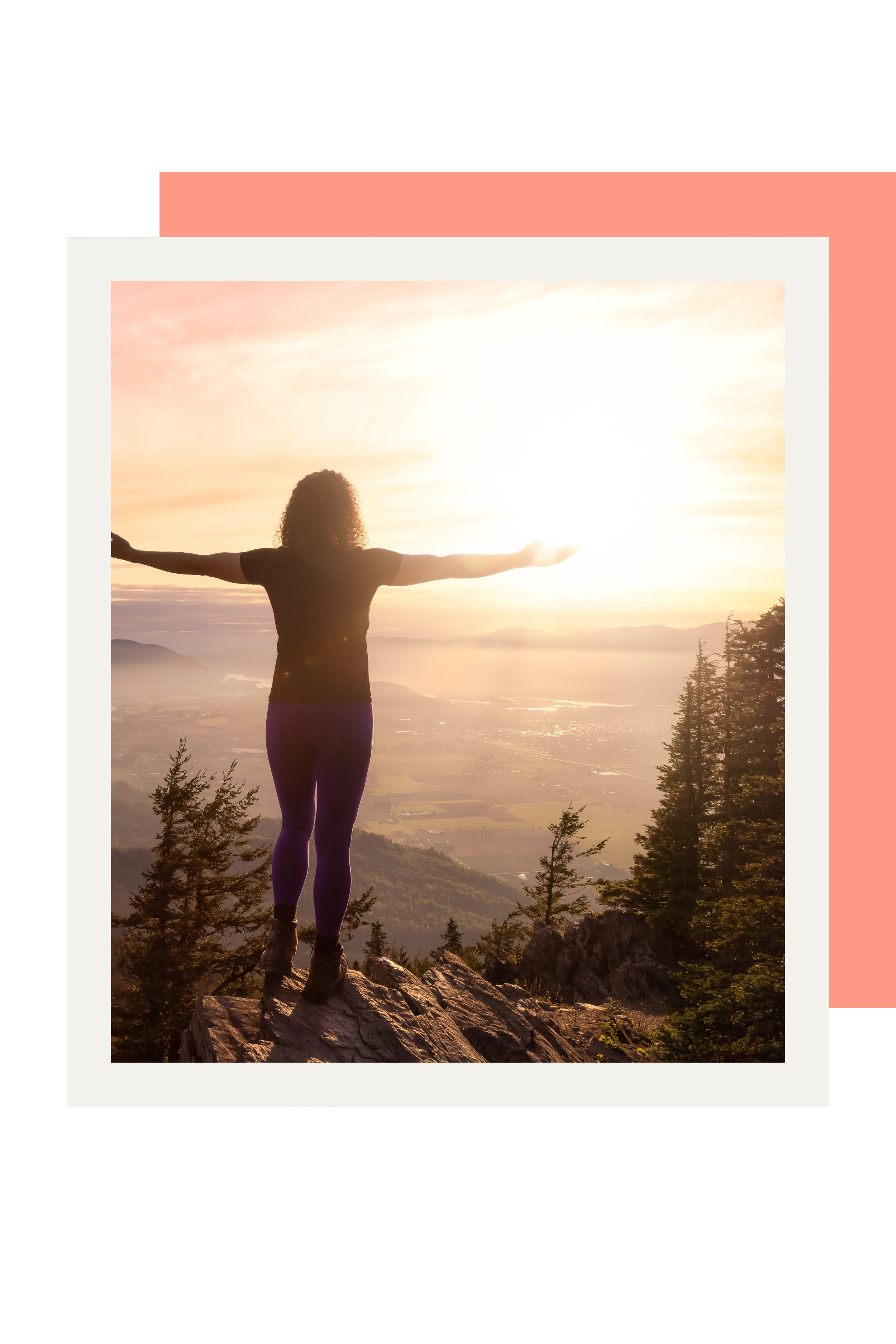 A woman with curly hair standing on a rocky ledge with arms outstretched, overlooking a valley with trees and mountains during sunrise or sunset.
