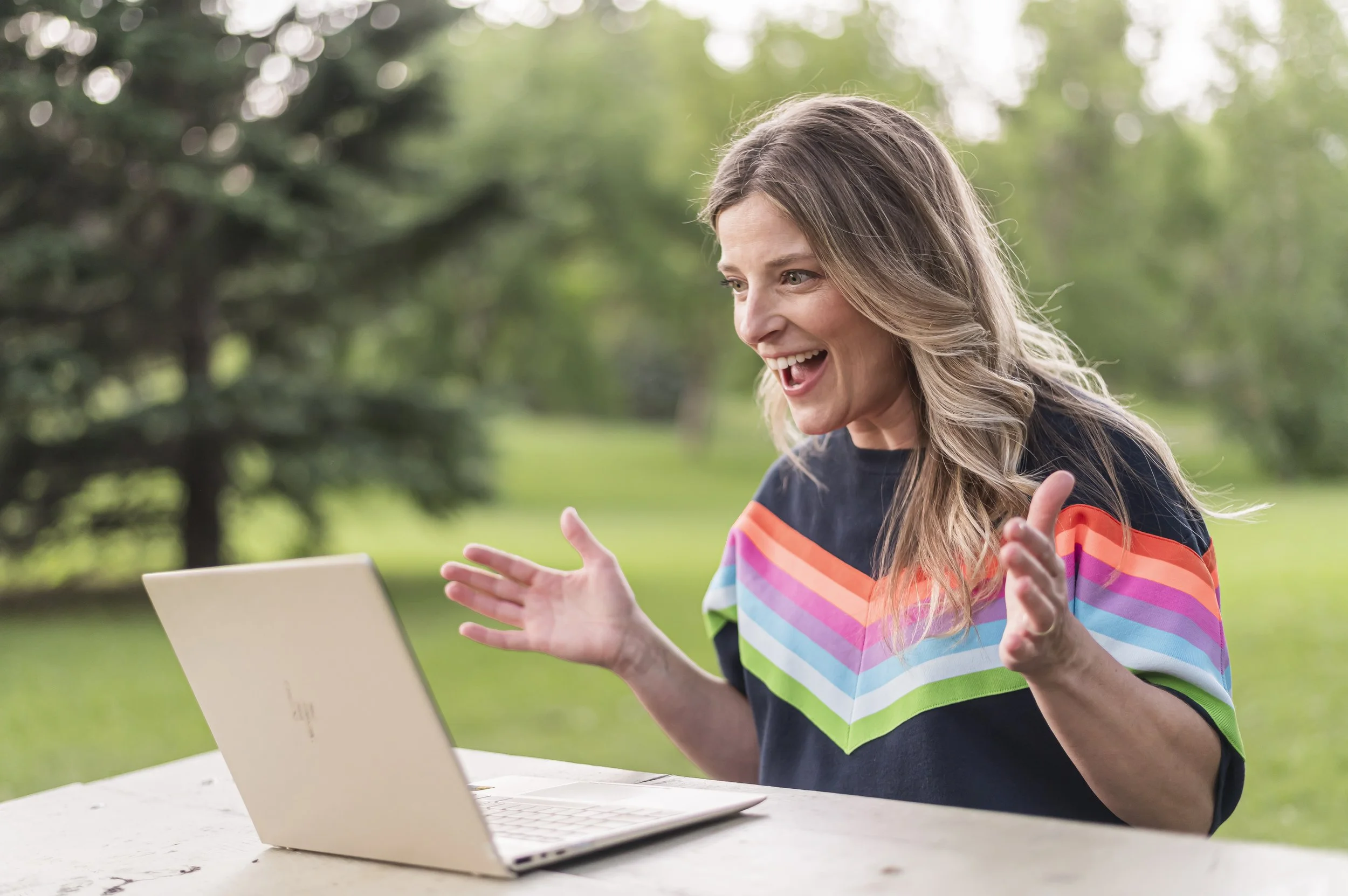 A woman enthusiastically talking during a video call outdoors with trees in the background.