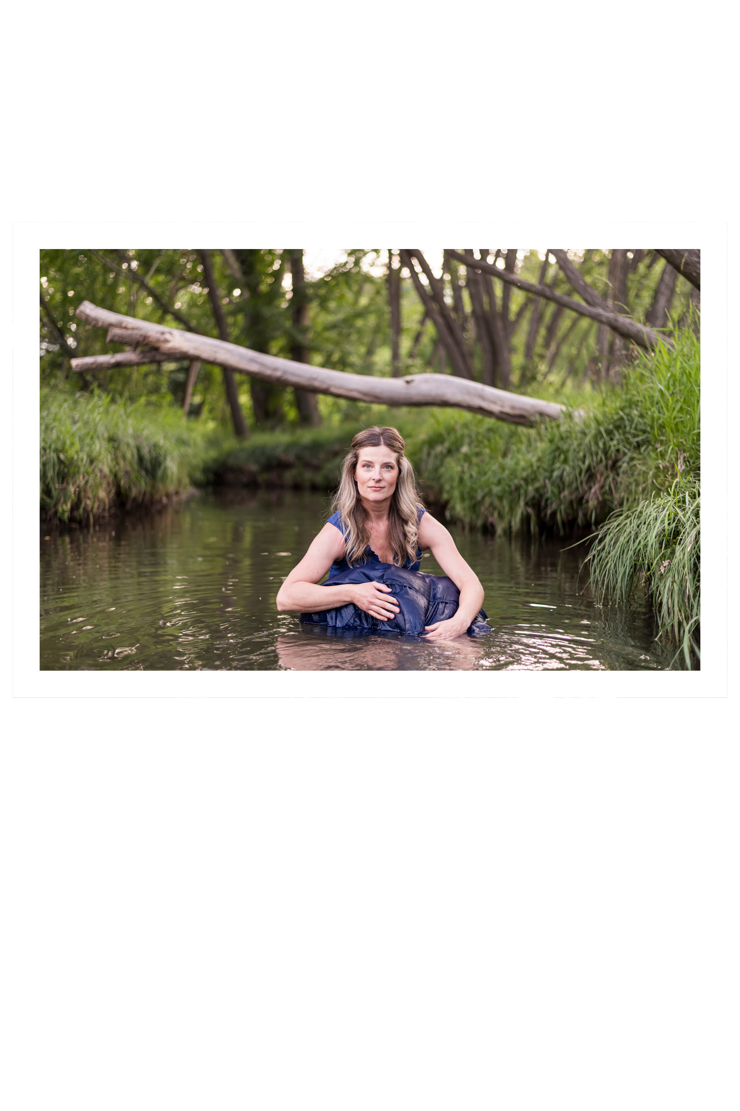 A woman with long wavy hair sitting waist-deep in a creek surrounded by greenery, with a fallen tree in the background.