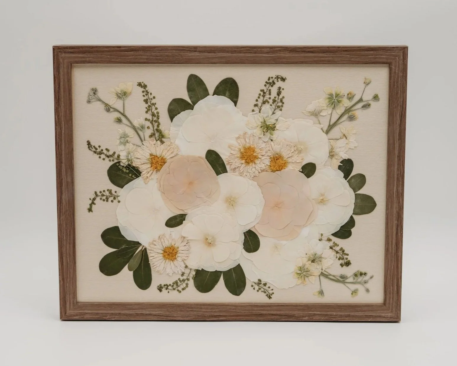 Framed pressed flower artwork with white, cream, and peach flowers, green leaves, and small yellow flowers on a beige background.