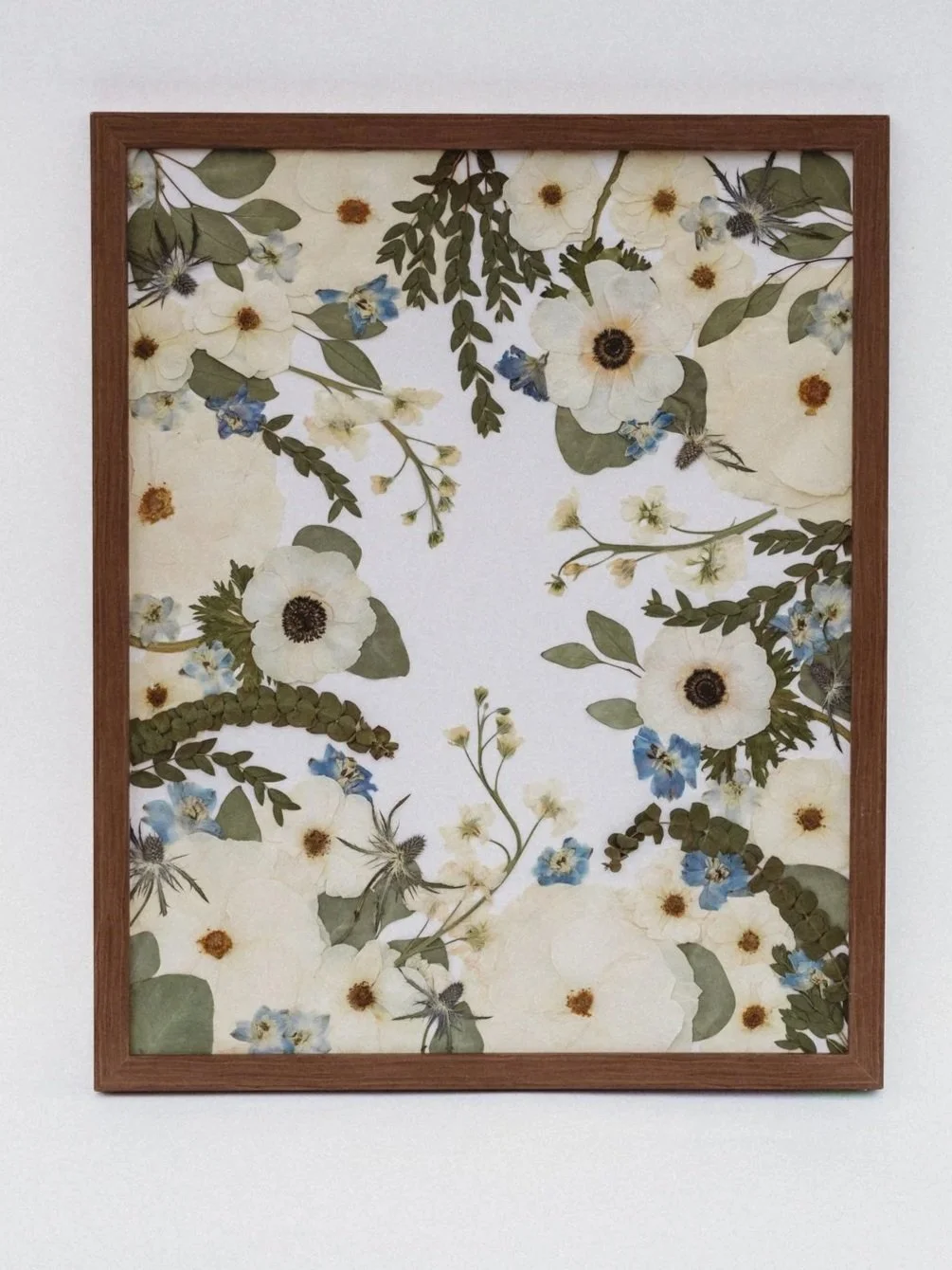 Framed artwork featuring a collage of pressed white, blue, and yellow flowers with green leaves on a white background.