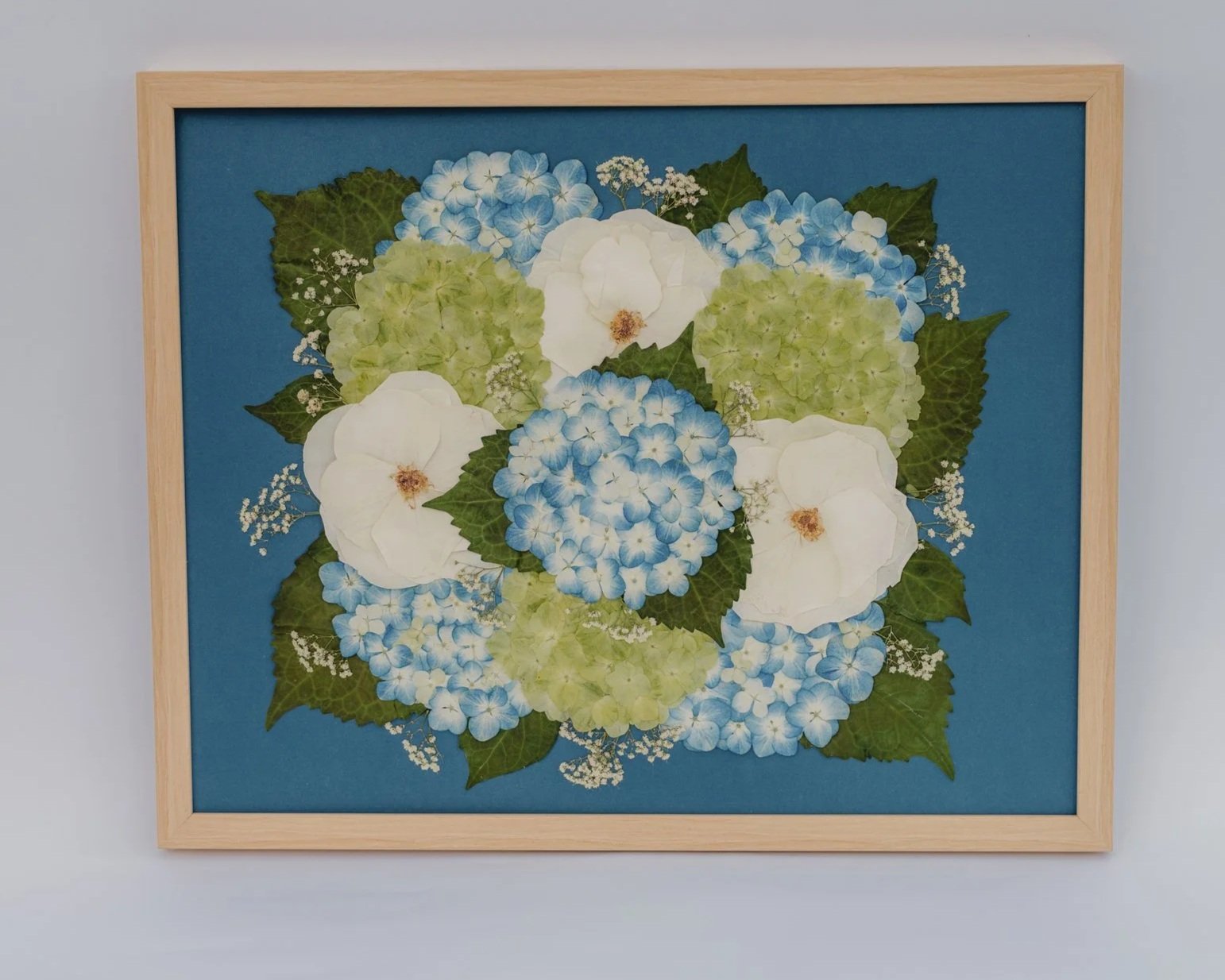 Framed artwork of a floral arrangement with white and blue hydrangeas and green leaves on a blue background.