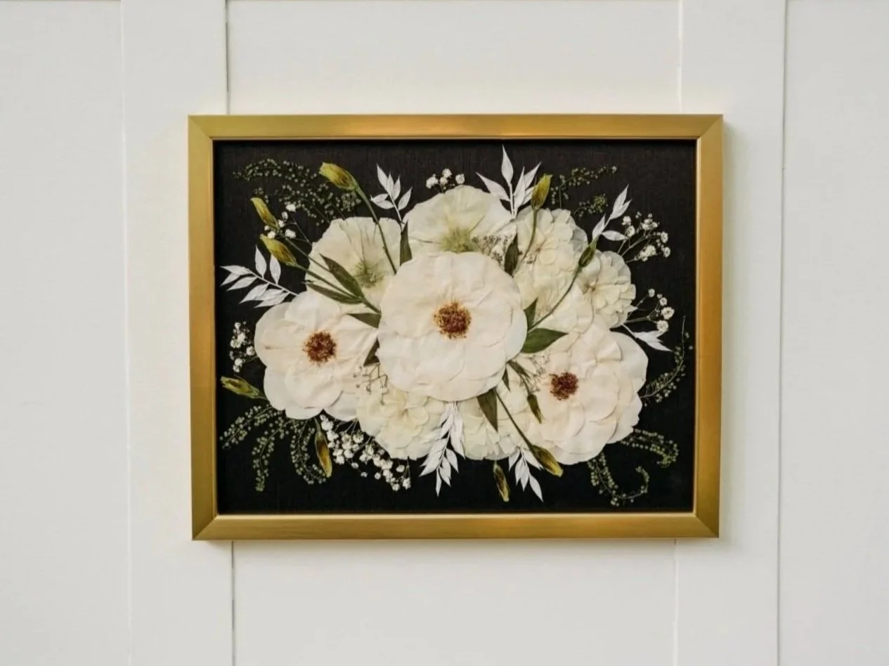Framed floral artwork featuring white flowers with brown centers and green leaves on a black background.