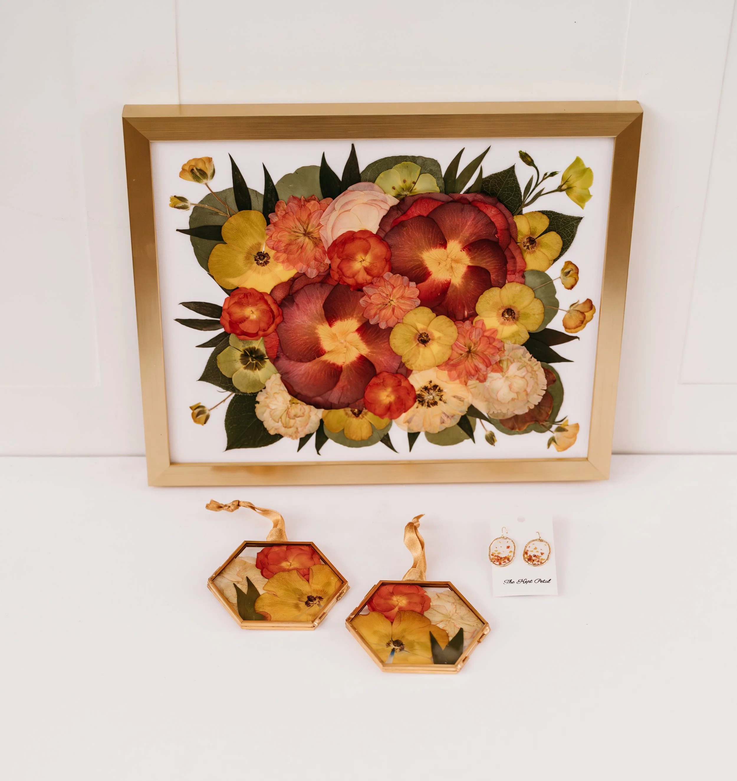 Decorative framed floral artwork with pressed flowers in a gold frame, matching pressed flower earrings, and a small card labeled 'The Key Petal' against a white background.