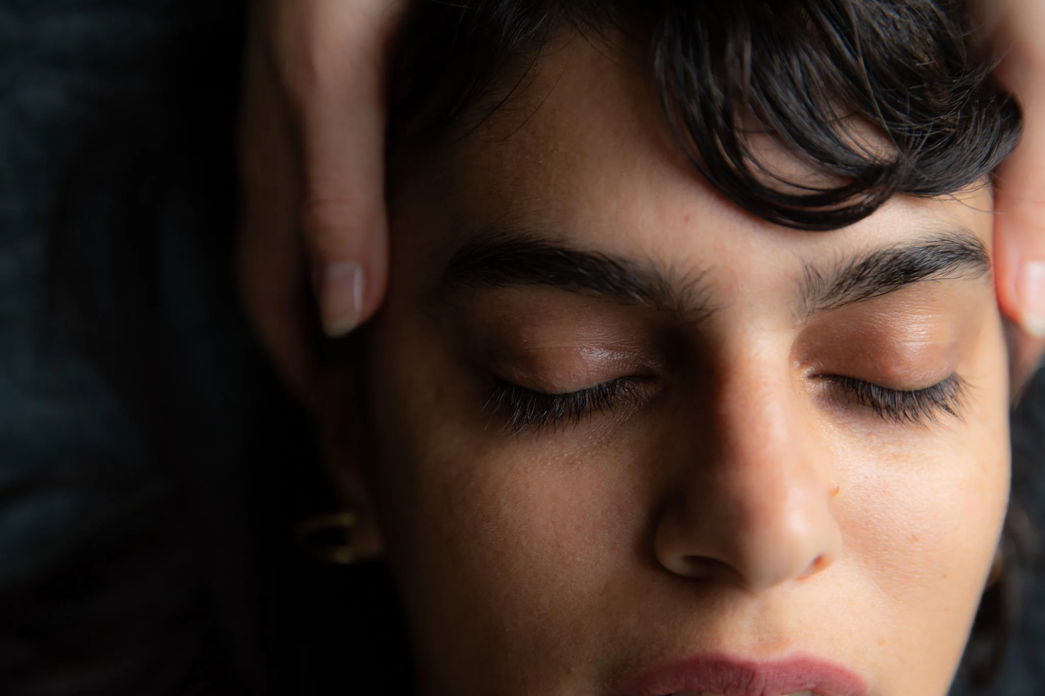 Close-up of a young woman's face with closed eyes, receiving a gentle head massage from someone with light skin.