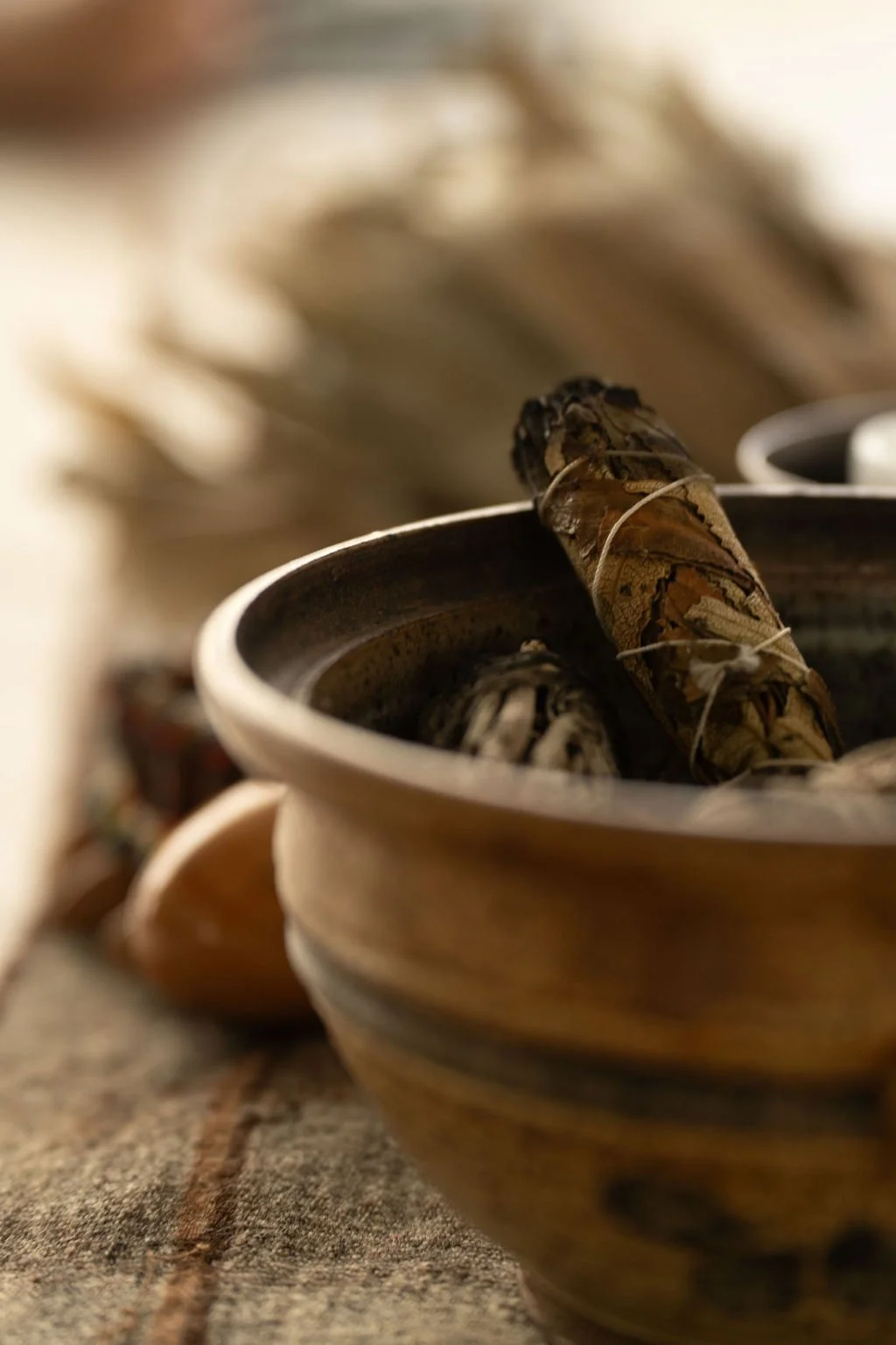 Close-up of a ceramic bowl containing wrapped smudge sticks, placed on a textured surface with a blurred background.