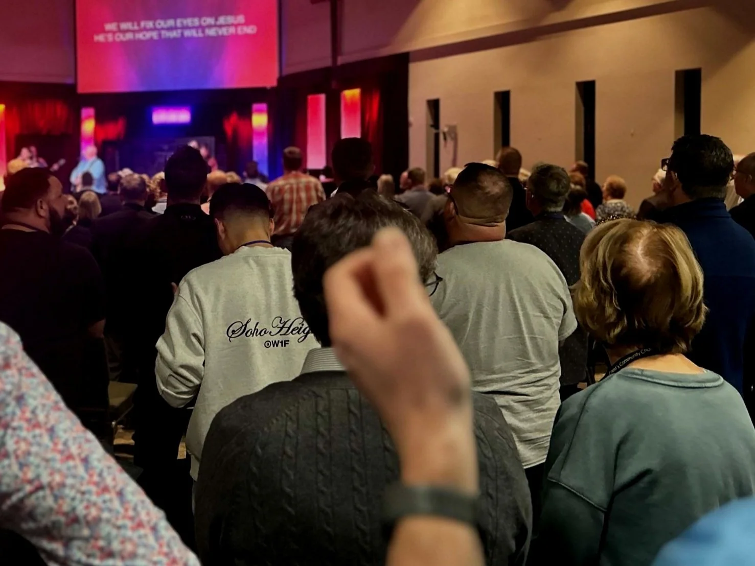 An open hymnal with sheet music on a black stand, positioned on a white table, next to a black pen, with a stained glass window in the background.