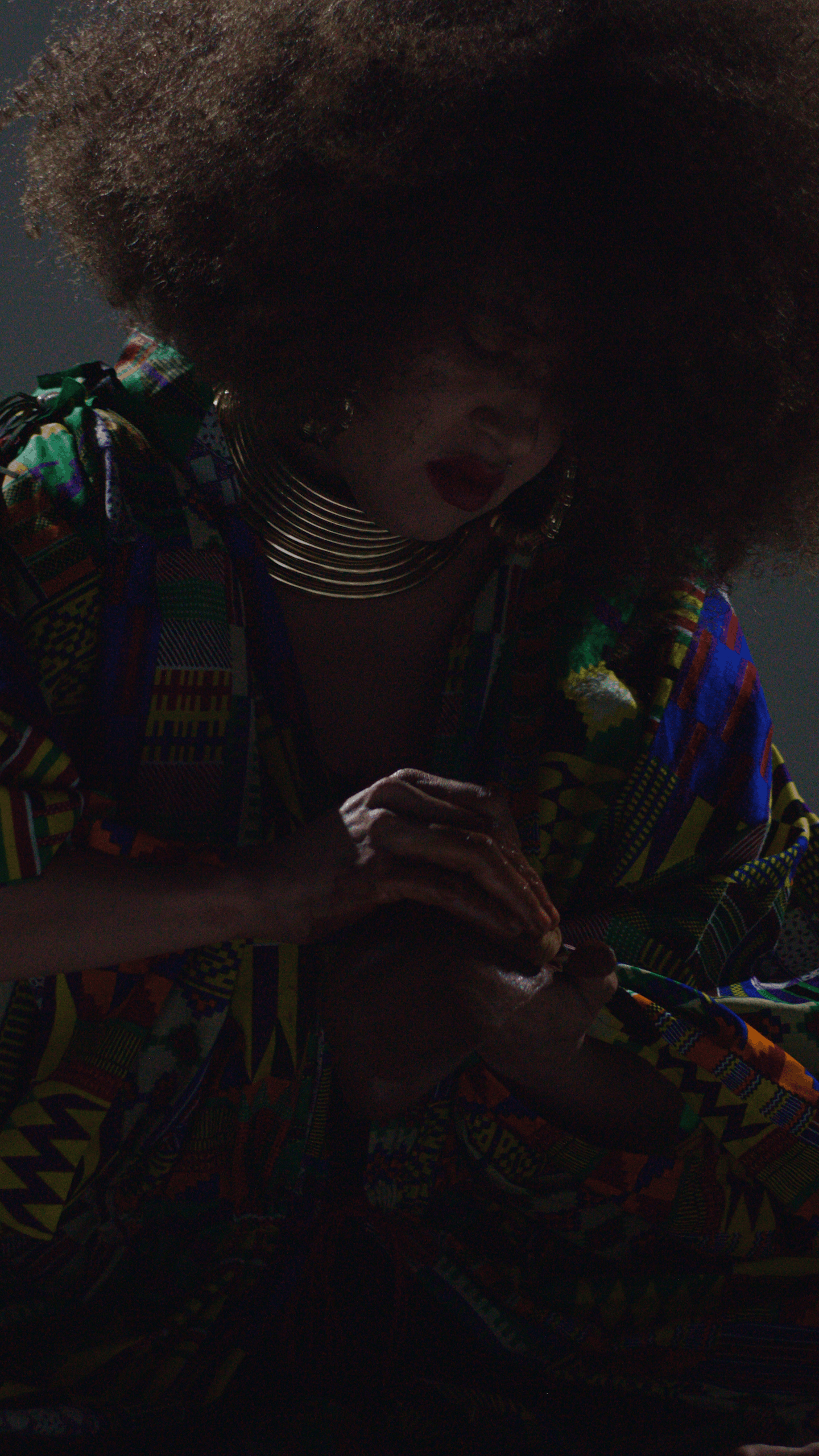 Woman wearing colorful traditional African clothing, large gold hoops, and a choker necklace, holding her hands together.