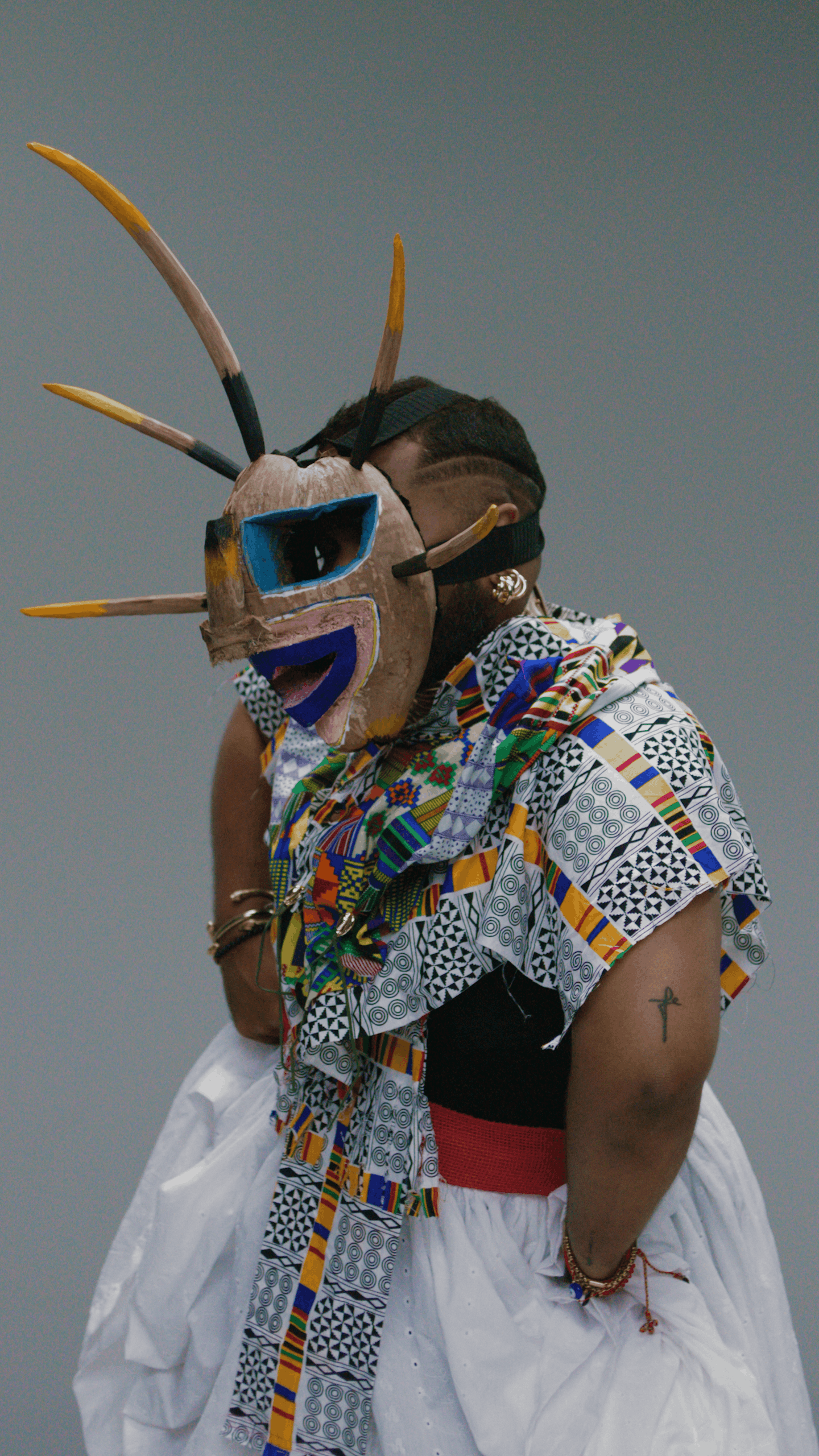 Person wearing a colorful patterned shirt and white skirt, with a vejigante mask resembling an animal face with exaggerated features and feathers, during a cultural performance or celebration.