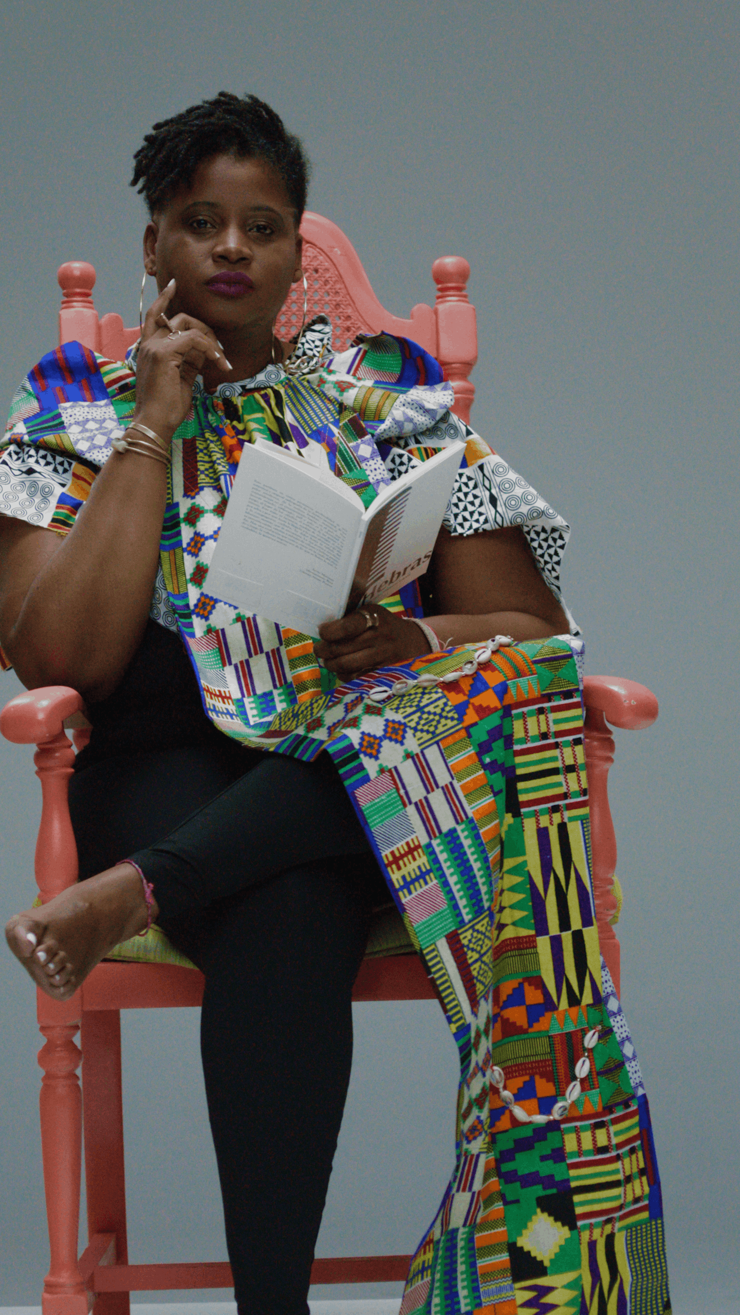 A woman sitting on a pink ornate wooden chair with a gray background. She is dressed in a colorful, patterned outfit, holding a book, and has short, natural hairstyle.