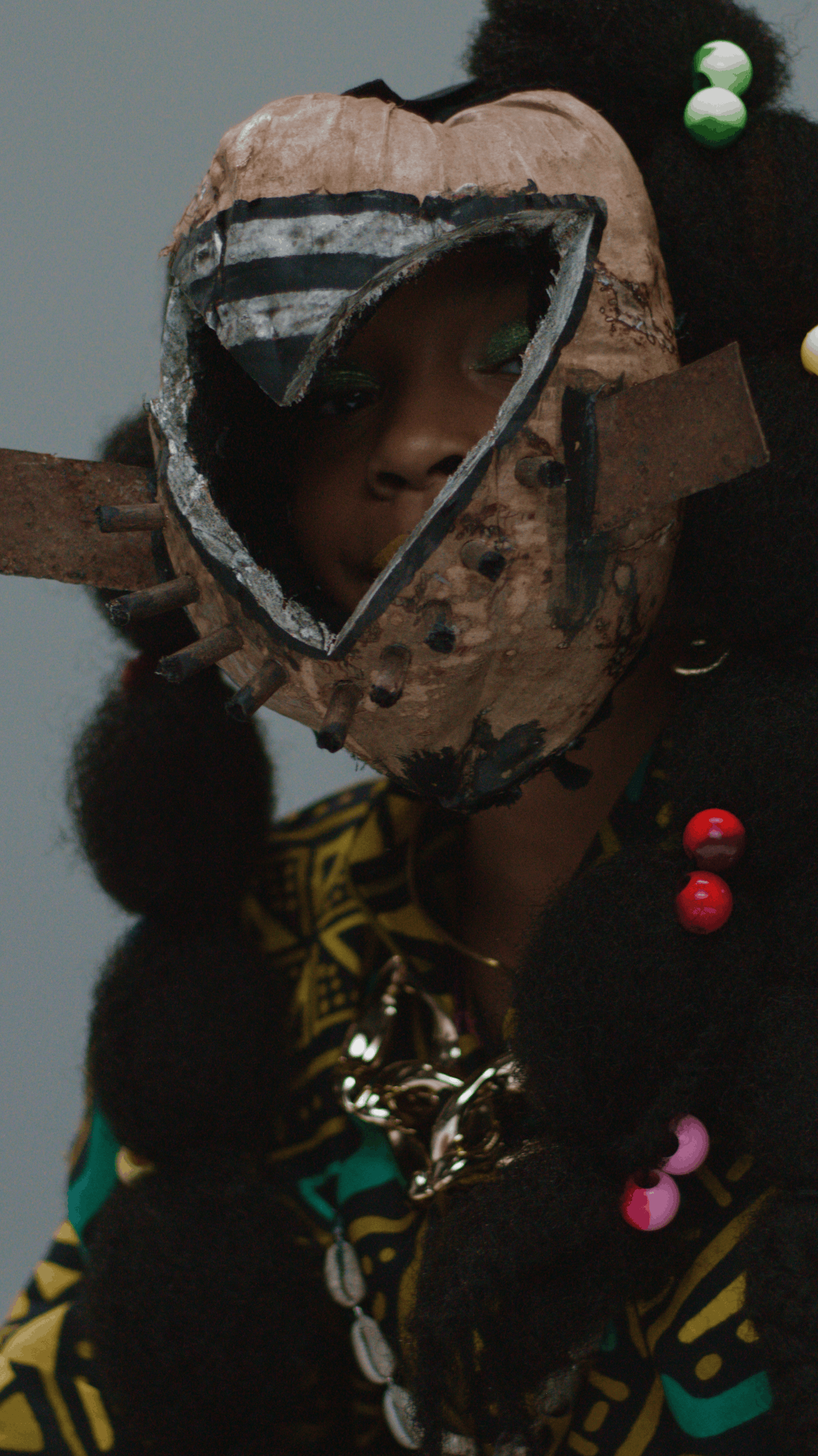 A person wearing a mask with a heart-shaped opening faces the camera. The mask appears handmade from metal and wood, with protruding nails or bolts. The person's eyes are visible through the eye holes, and they have vibrant green eye makeup. The indi