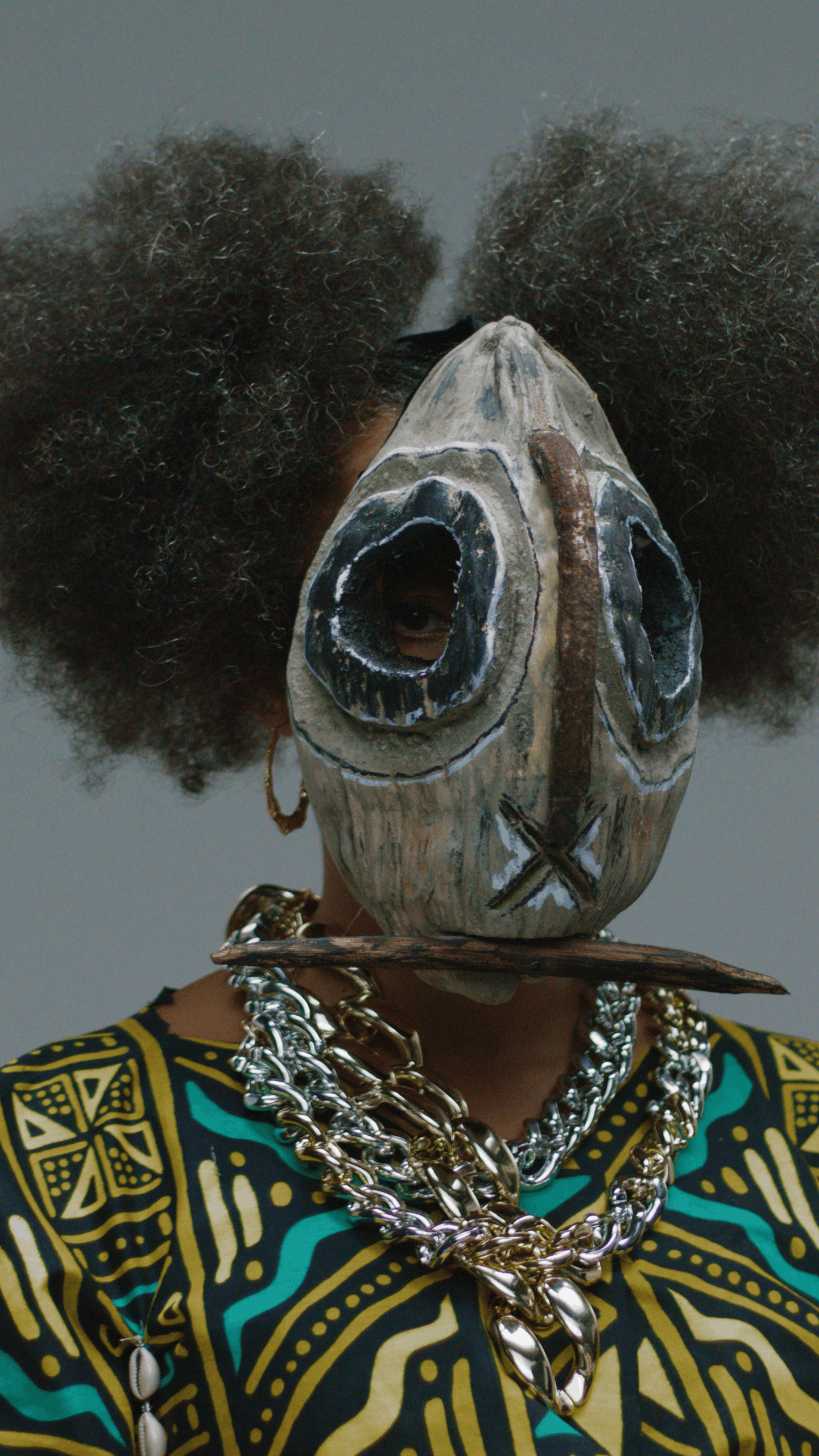 A person wearing a vejigante mask with eye holes, curly hair styled in two puffs, gold earrings, and layered silver necklaces, dressed in a colorful, patterned top.