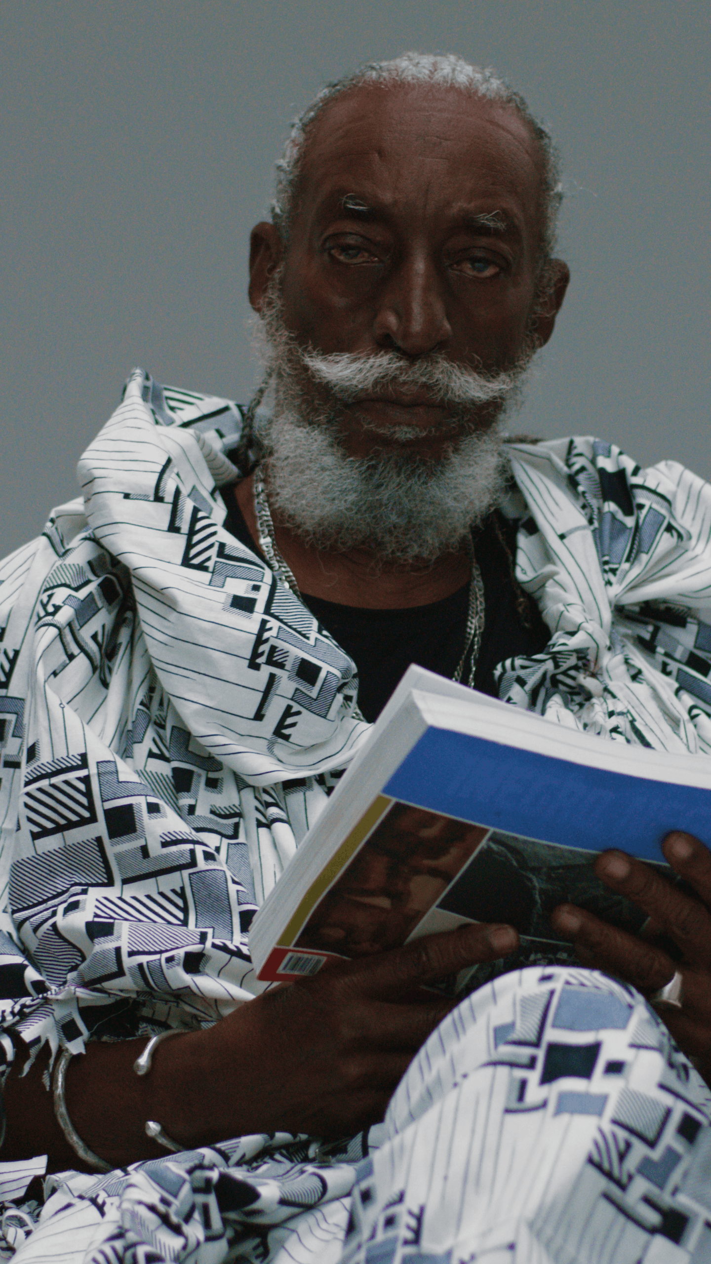 An elderly man with gray hair and beard, wearing a patterned black-and-white outfit, sitting and reading a book.