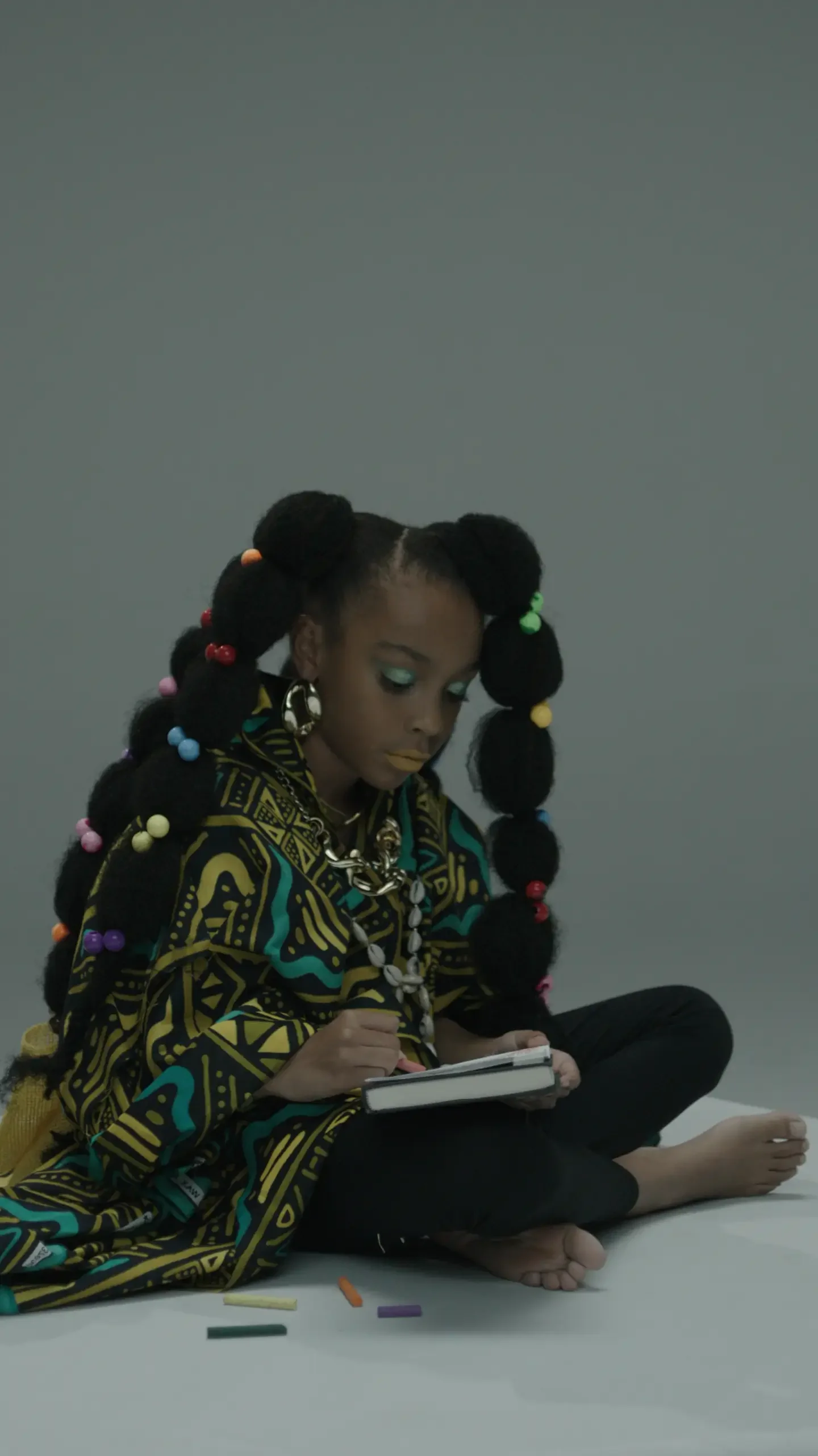 A girl with elaborate hair styled in large, colorful beads, wearing vibrant makeup, a patterned shirt, and chunky jewelry, sitting barefoot on the floor using a notepad and pen.