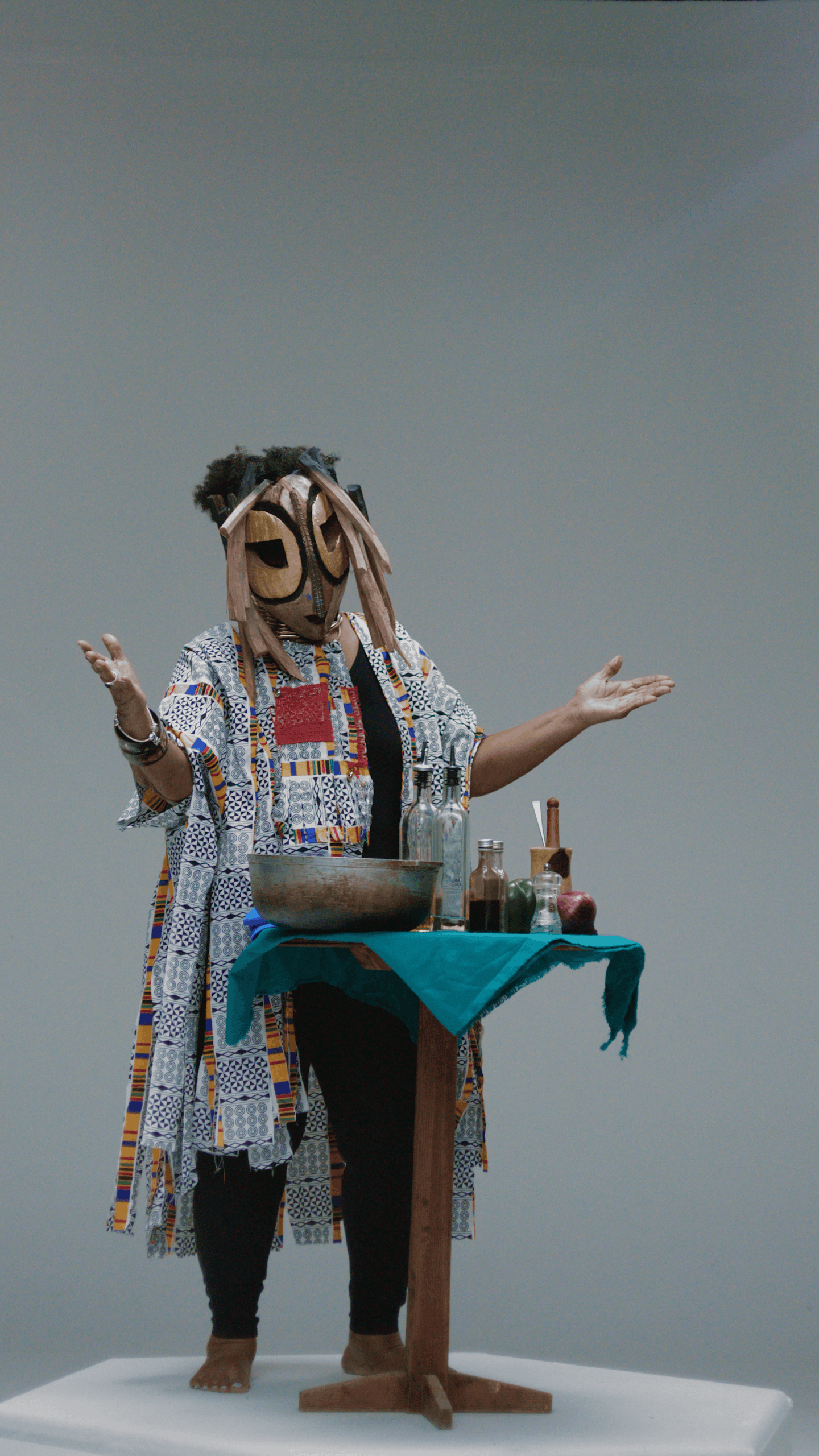 Person wearing a large coconut mask, standing behind a table with potion bottles, a bowl, and other items, gesturing with open arms.
