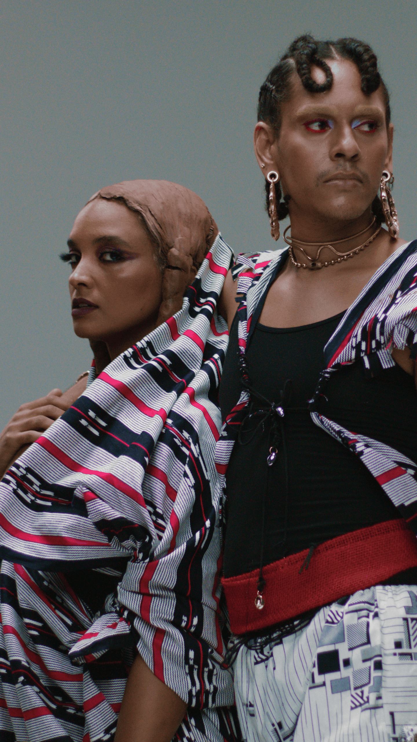 Two individuals standing closely, both dressed in colorful, patterned clothing, with serious expressions, against a plain background.