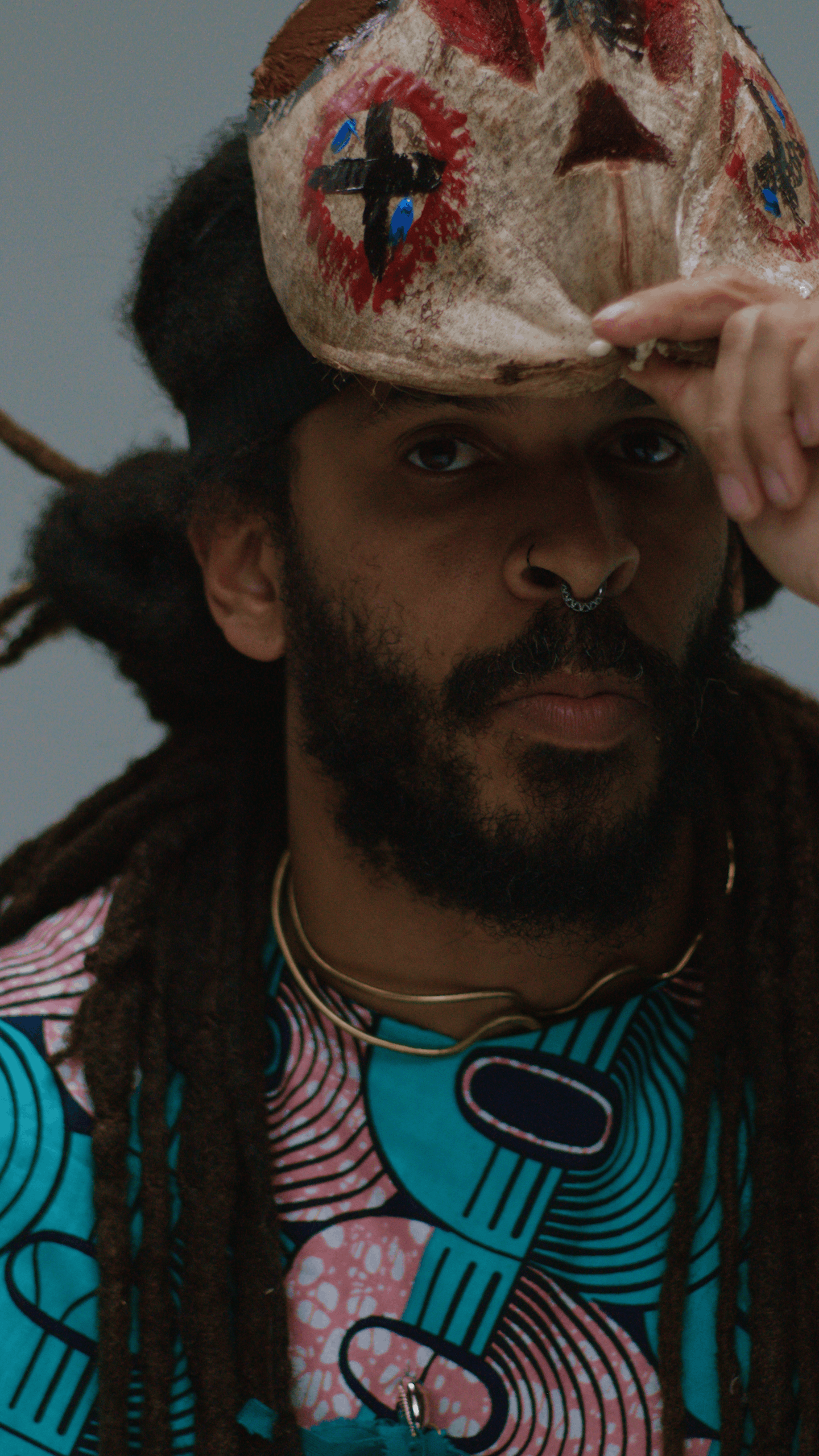 A man with dreadlocks, a nose ring, and wearing a vibrant, patterned shirt holds a painted vejigante mask above his head.