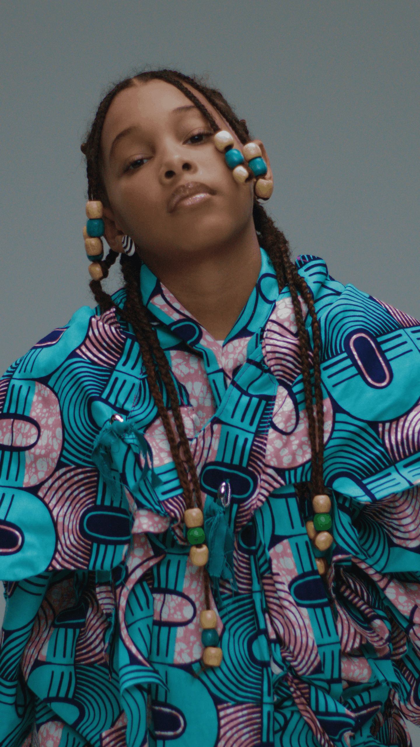 A child with styled beads in her braids, wearing a colorful patterned jacket and necklace, posing with a serious expression.