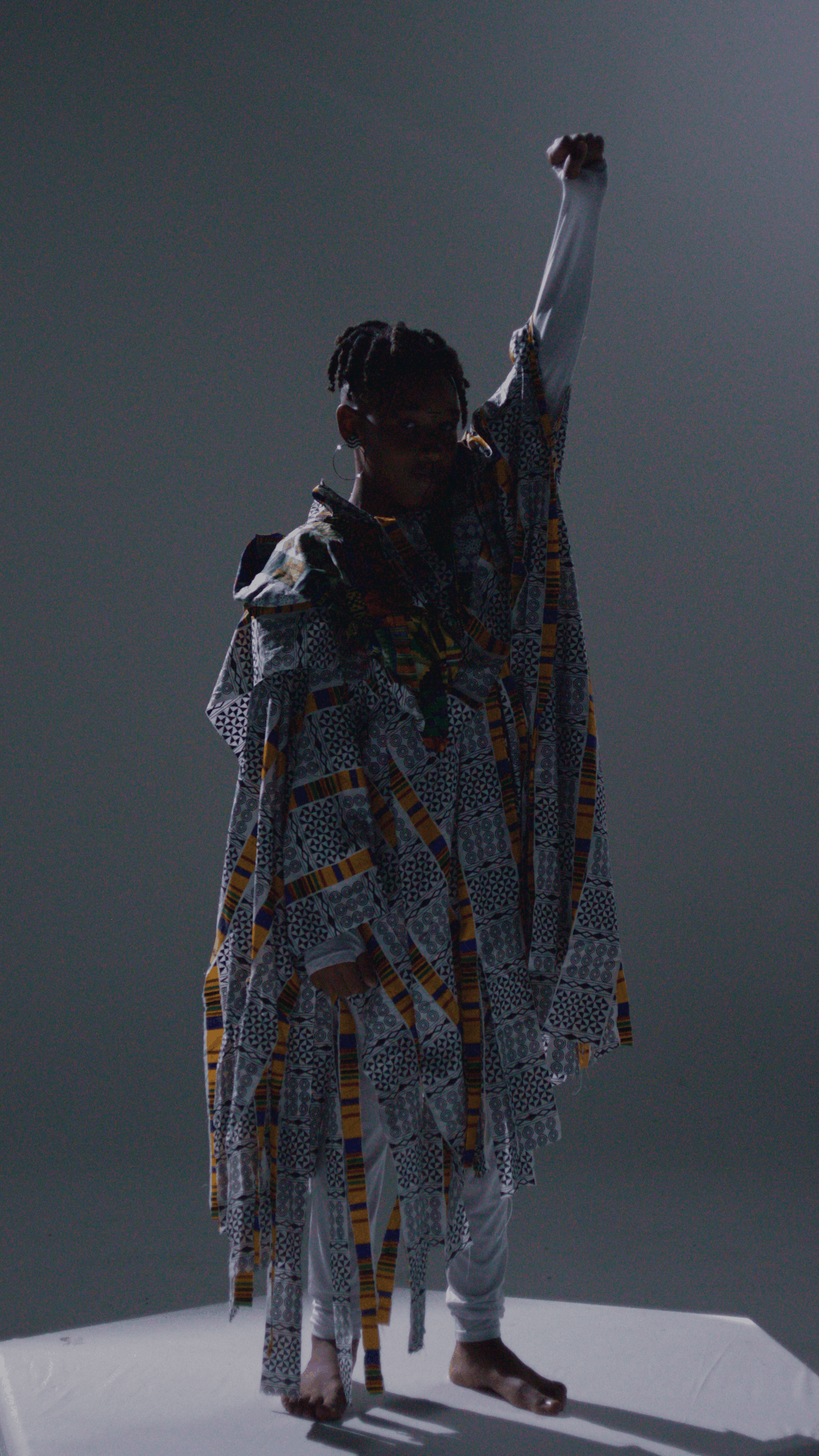 A kid with braided hair dressed in a patterned, multi-colored kente cloth, standing barefoot on a white surface against a plain grey background, with one arm raised.