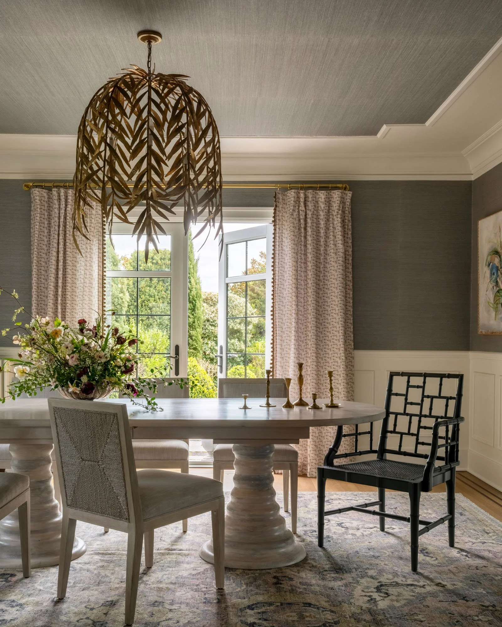 A dining room with a round white table, four chairs, a flower arrangement, hanging leafy chandelier, and open glass doors with beige curtains leading to a green garden.