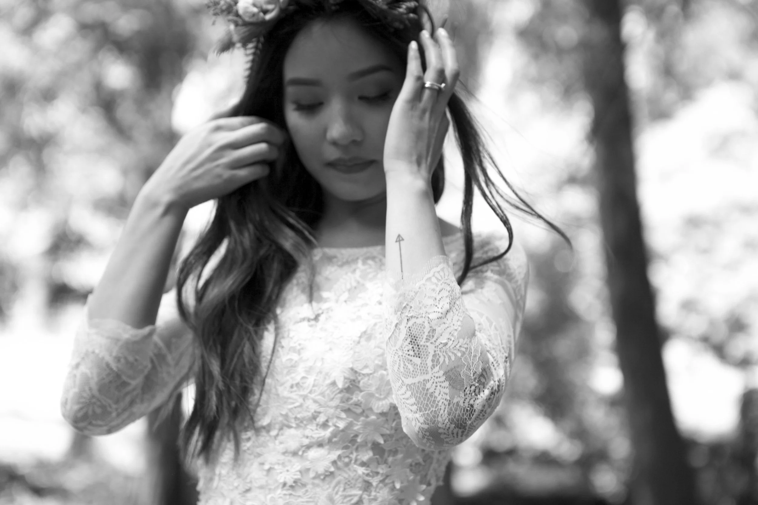 Woman in lace dress with floral headband and arrow tattoo on forearm, touching her hair.