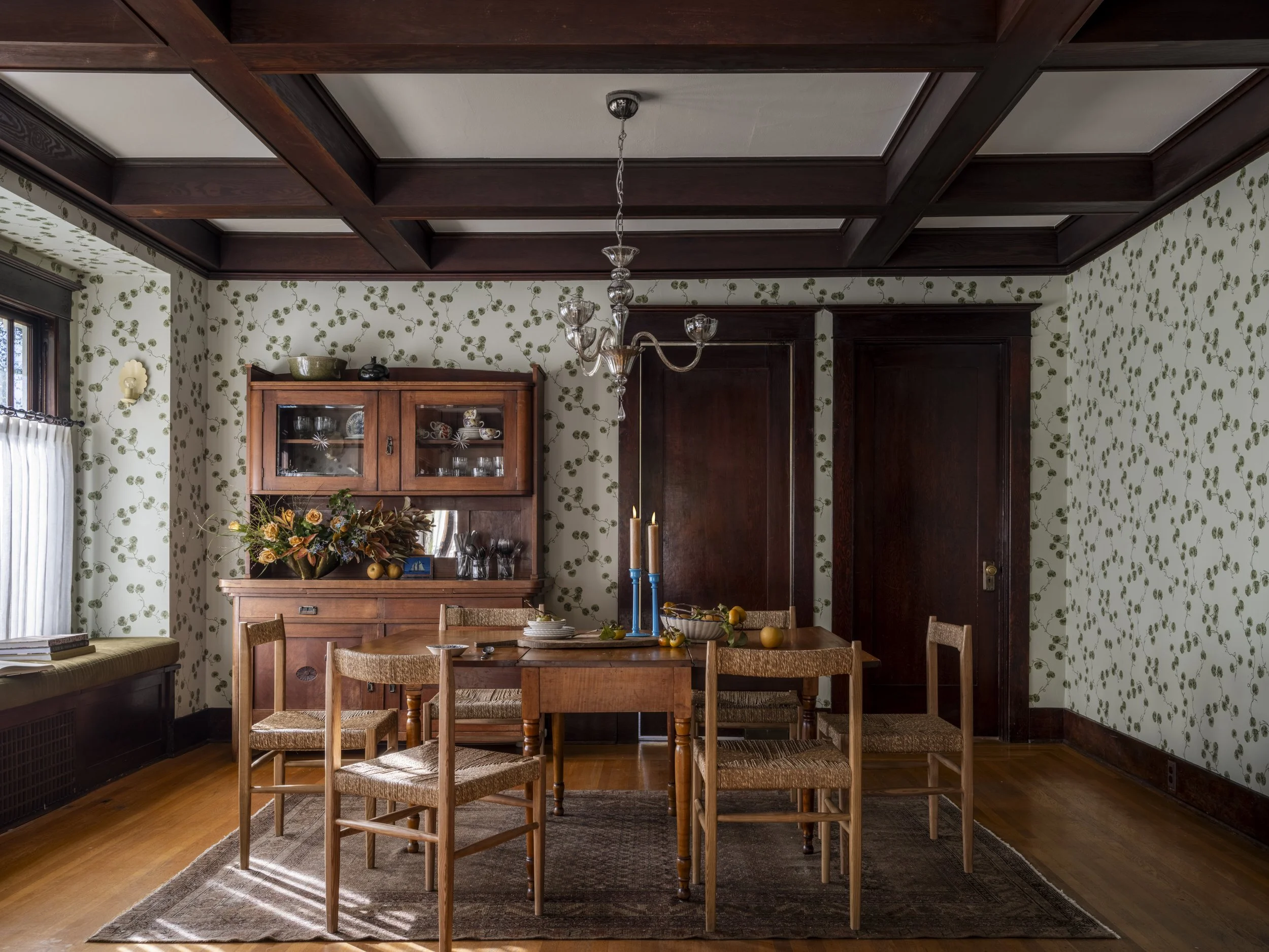 Vintage dining room with wooden furniture, patterned wallpaper, and a chandelier. A wooden dining table is surrounded by woven chairs, and a sideboard displays decorative items. The room has a cozy, retro style.
