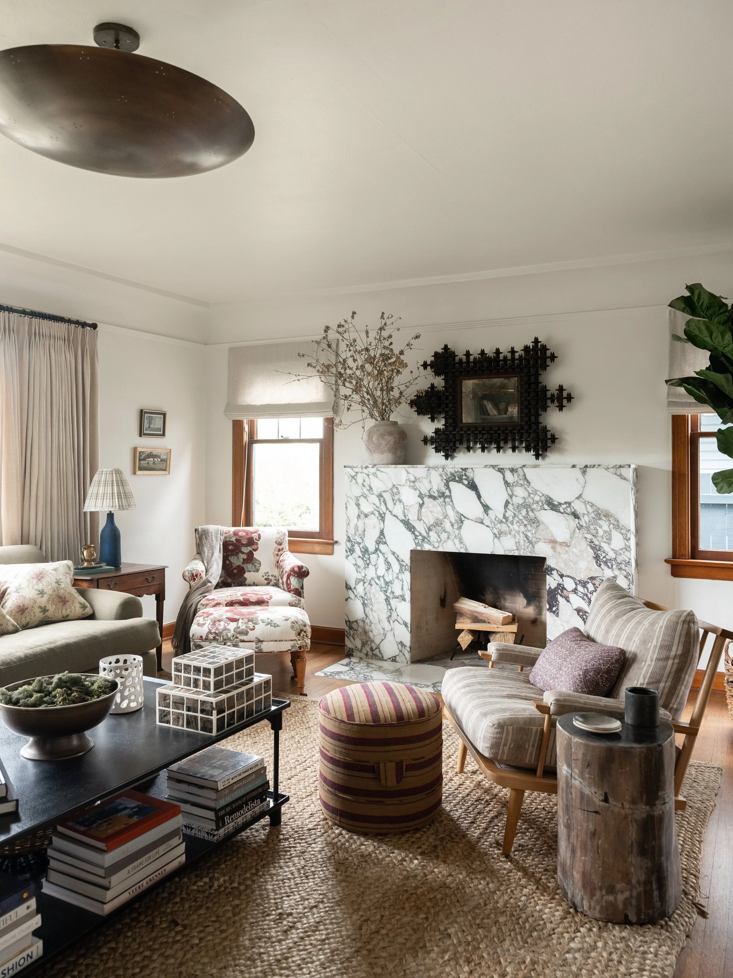 cozy living room with eclectic decor, featuring a marble fireplace, patterned armchair and footstool, woven rug, and books stacked on a coffee table