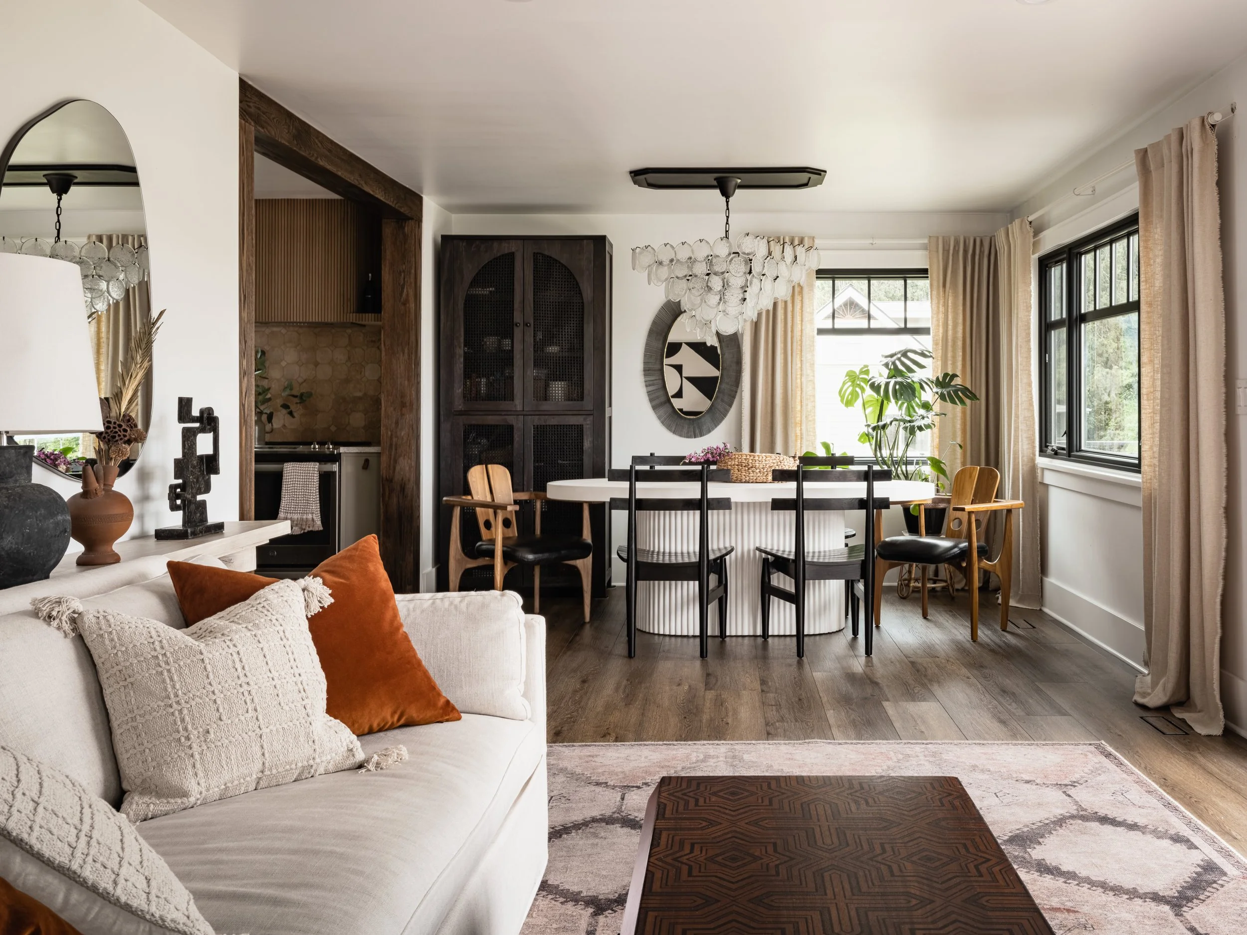 Modern living and dining room with beige sofa, orange and cream pillows, round dining table with black chairs, chandelier, large windows with curtains, decorative plants, wooden cabinet, and artistic decor.