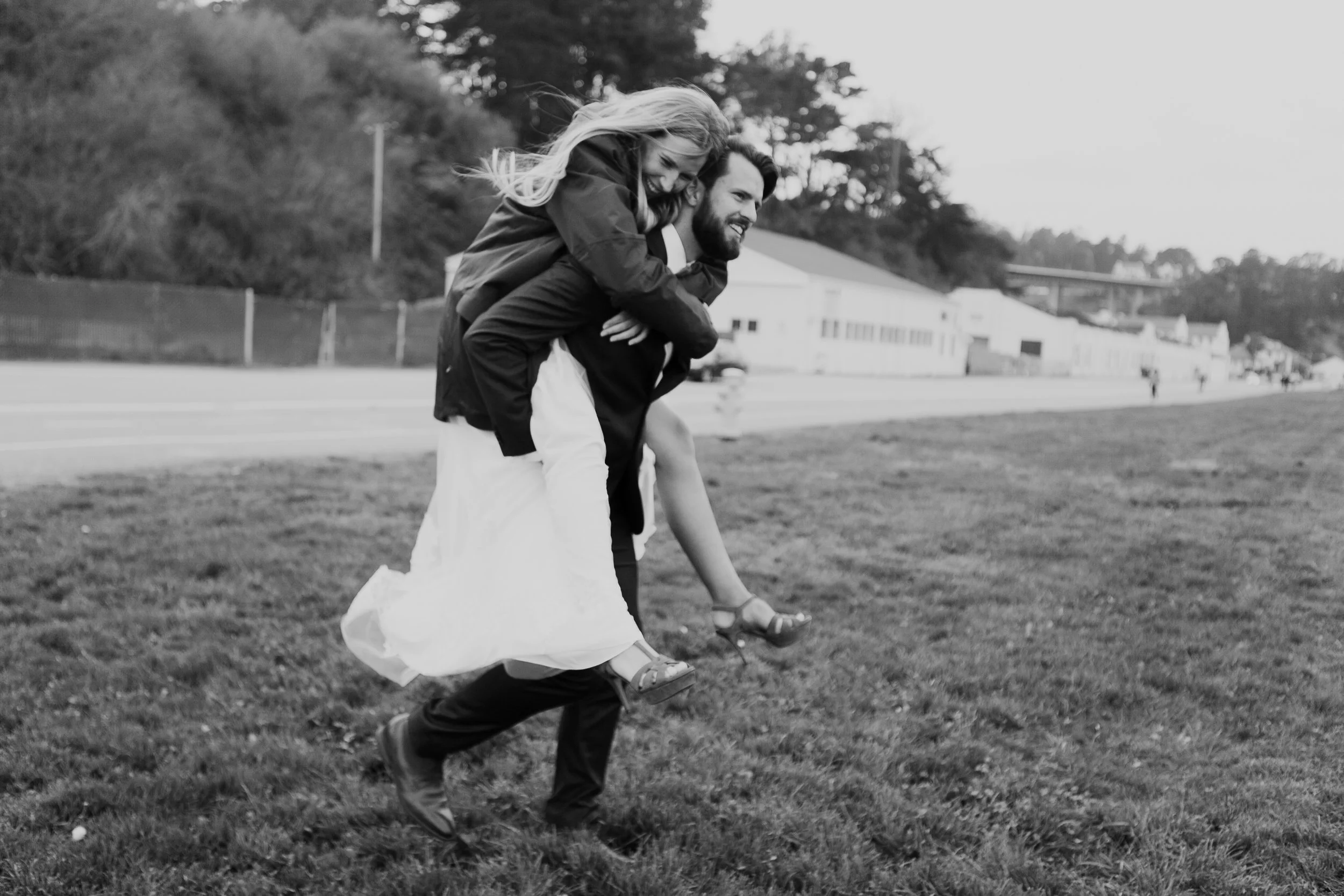 Black and white photo of a man in a suit giving a woman a piggyback ride. The woman is wearing a dress and smiling, with a background of grass, a road, and trees.