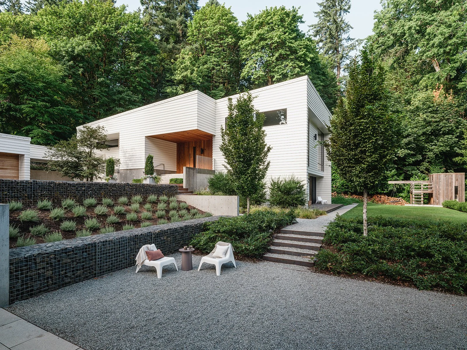 Modern white house surrounded by lush greenery, featuring a tiered garden with shrubs and steps. Two white chairs with a small table are placed on a gravel path in the foreground.