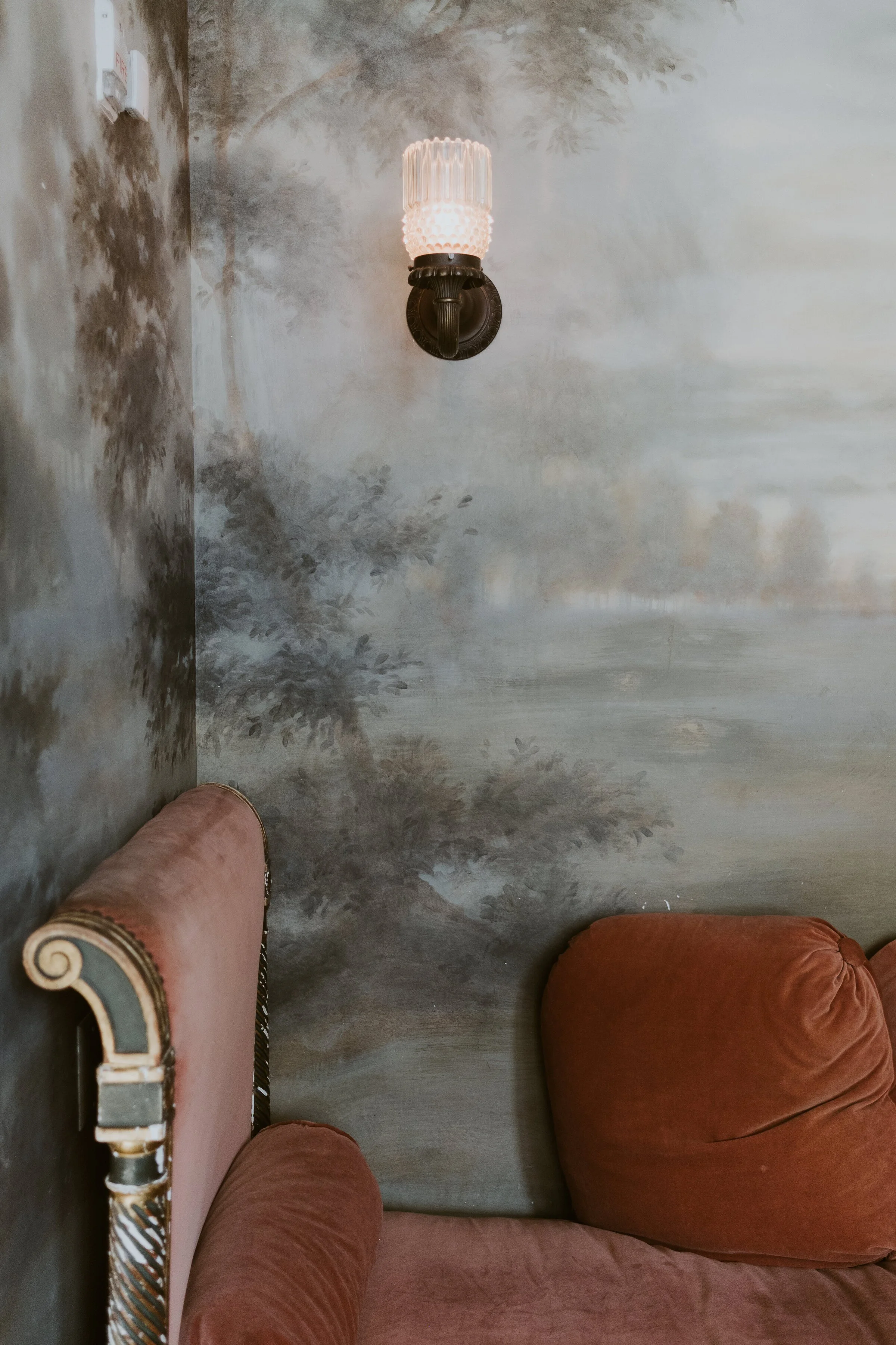 Vintage room corner with a wall sconce light, a landscape mural, and a retro velvet couch.