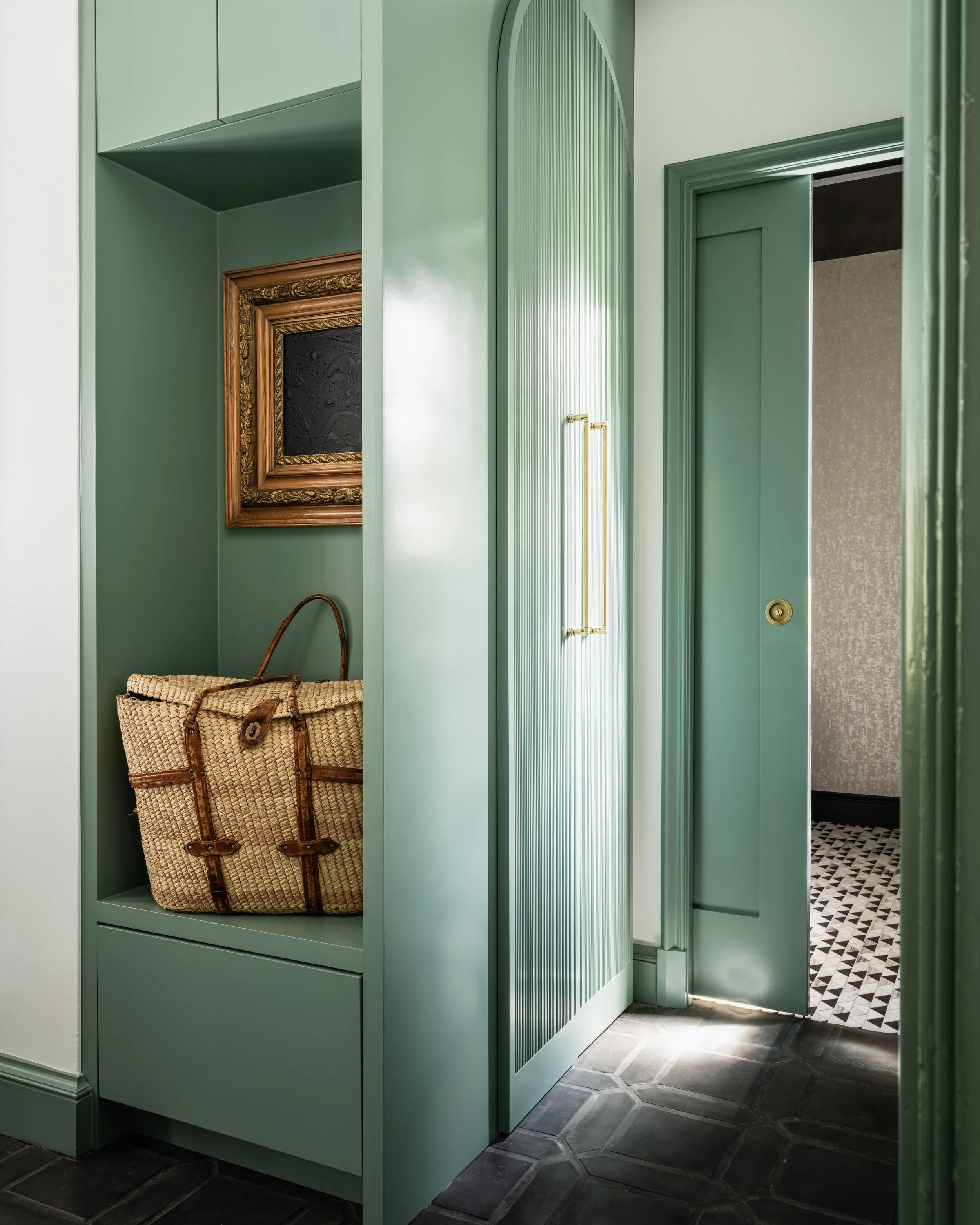 Green built-in storage unit with woven basket and framed artwork in a hallway with geometric floor tiles.