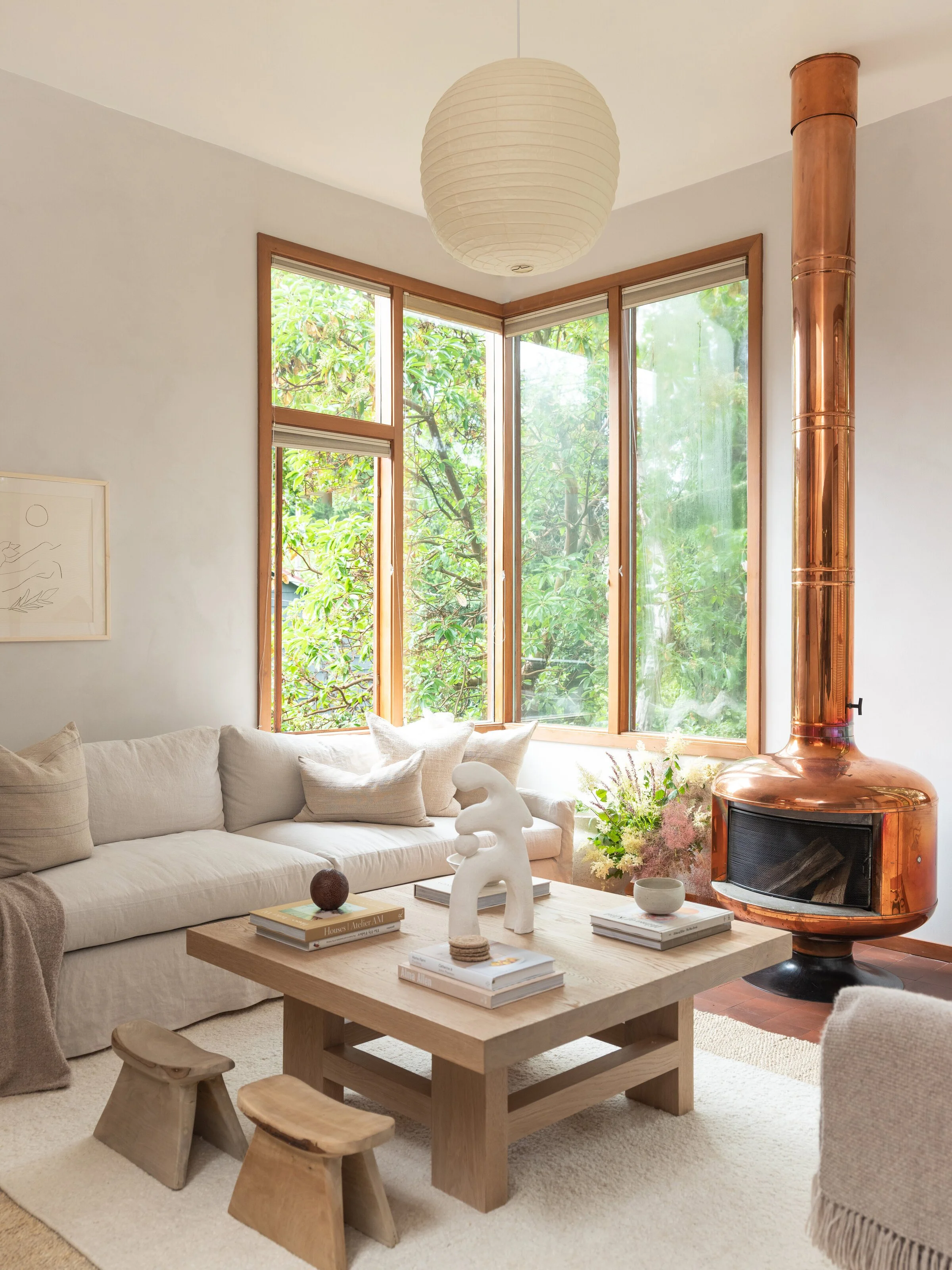 Modern living room with a beige sectional sofa, wooden coffee table with decorative items, two small wooden stools, copper fireplace, large windows with greenery outside, and a paper lantern pendant light.