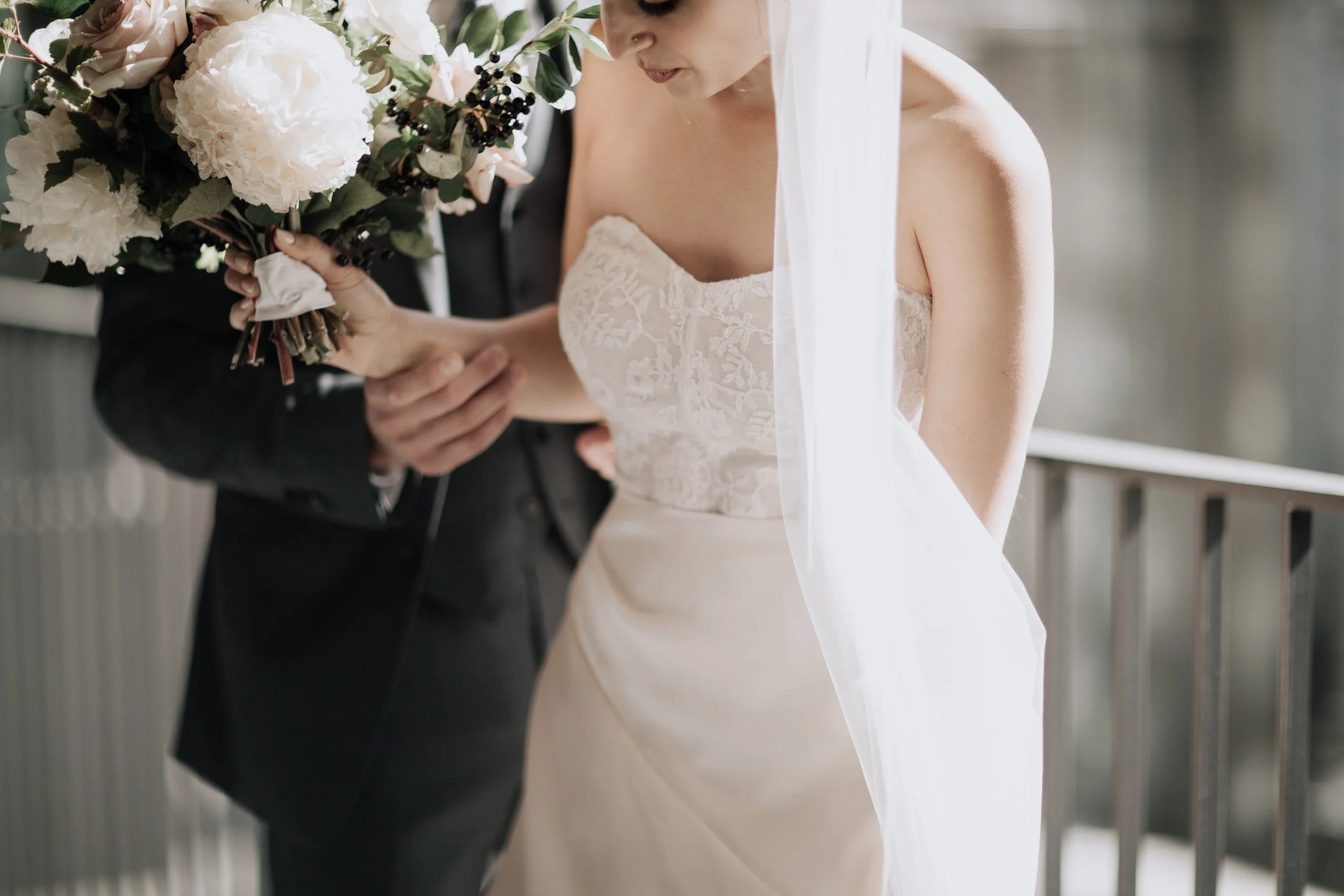 Bride and groom holding a bouquet of white and pink flowers, with bride wearing a lace wedding dress and veil, groom in a dark suit.