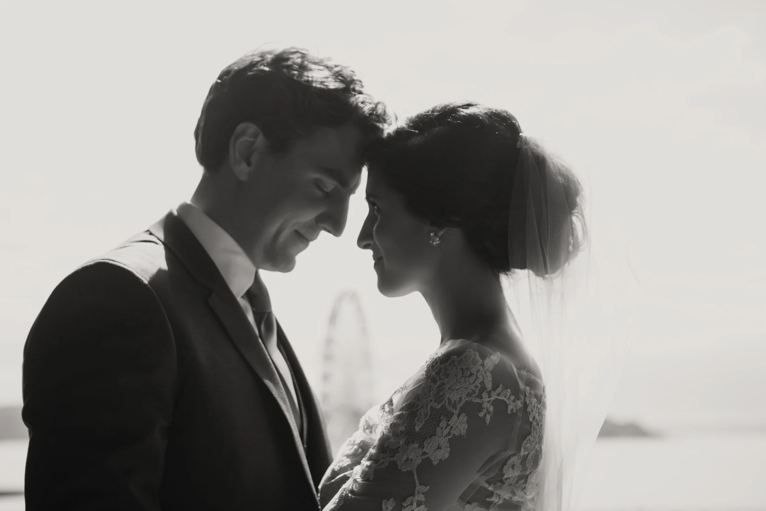Black and white photo of a bride and groom embracing, smiling, with a Ferris wheel in the background.