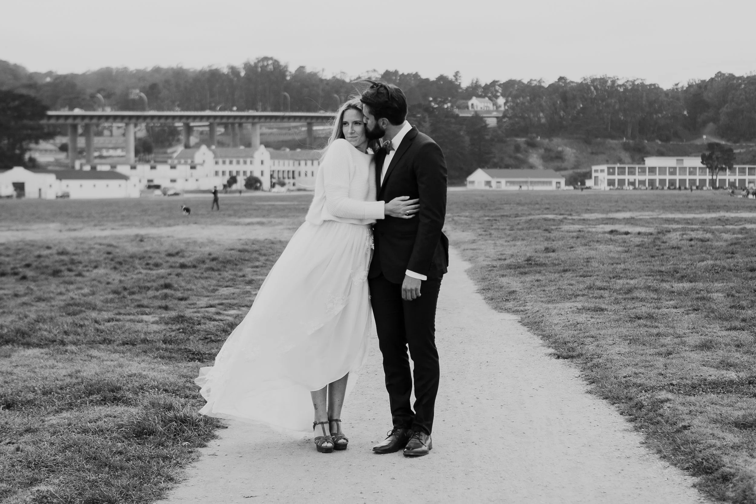Bride and groom standing on a path in a park, black and white photo, wearing wedding attire, with trees and buildings in the background.