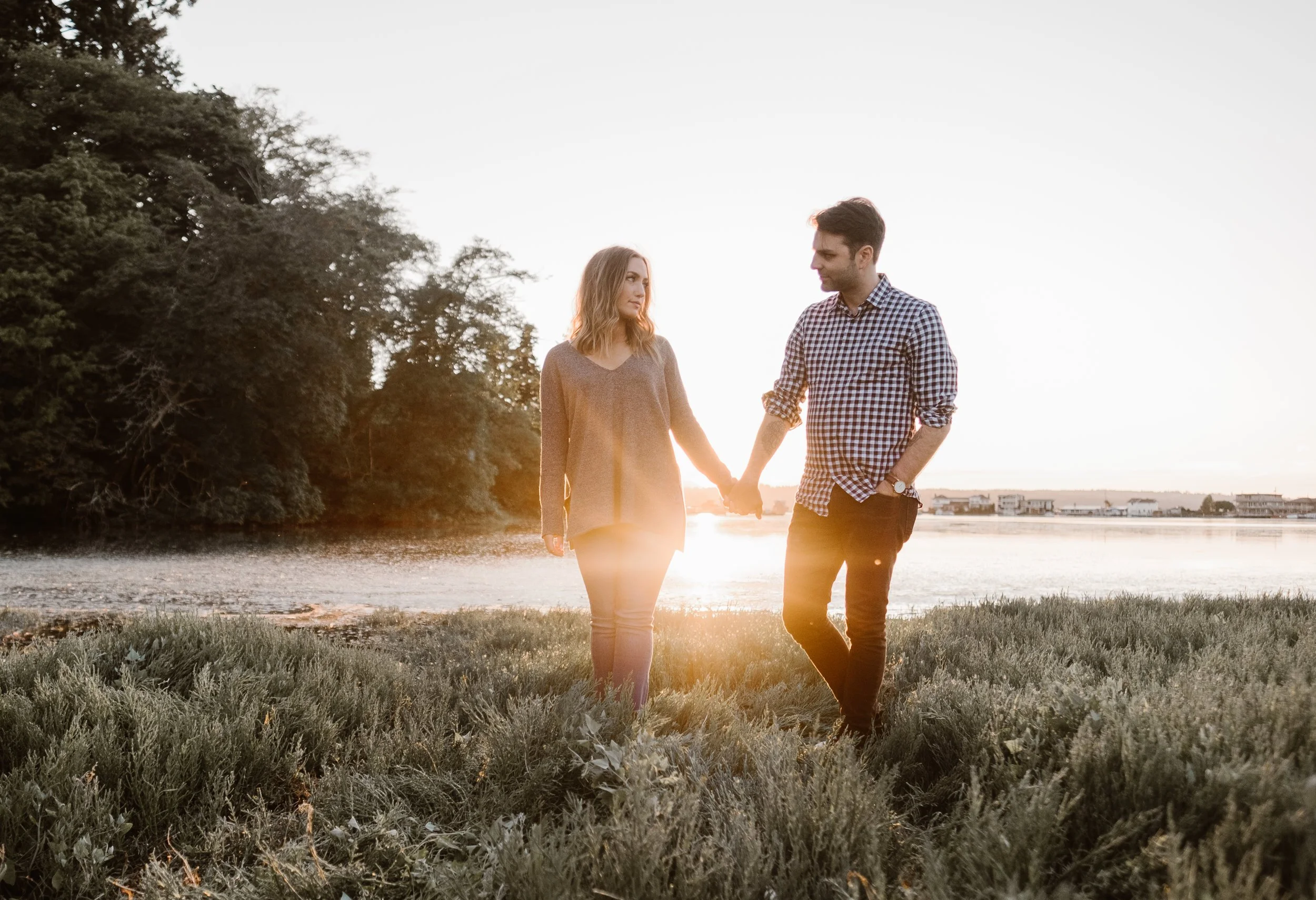 Couple holding hands near a lake at sunset with trees in the background.